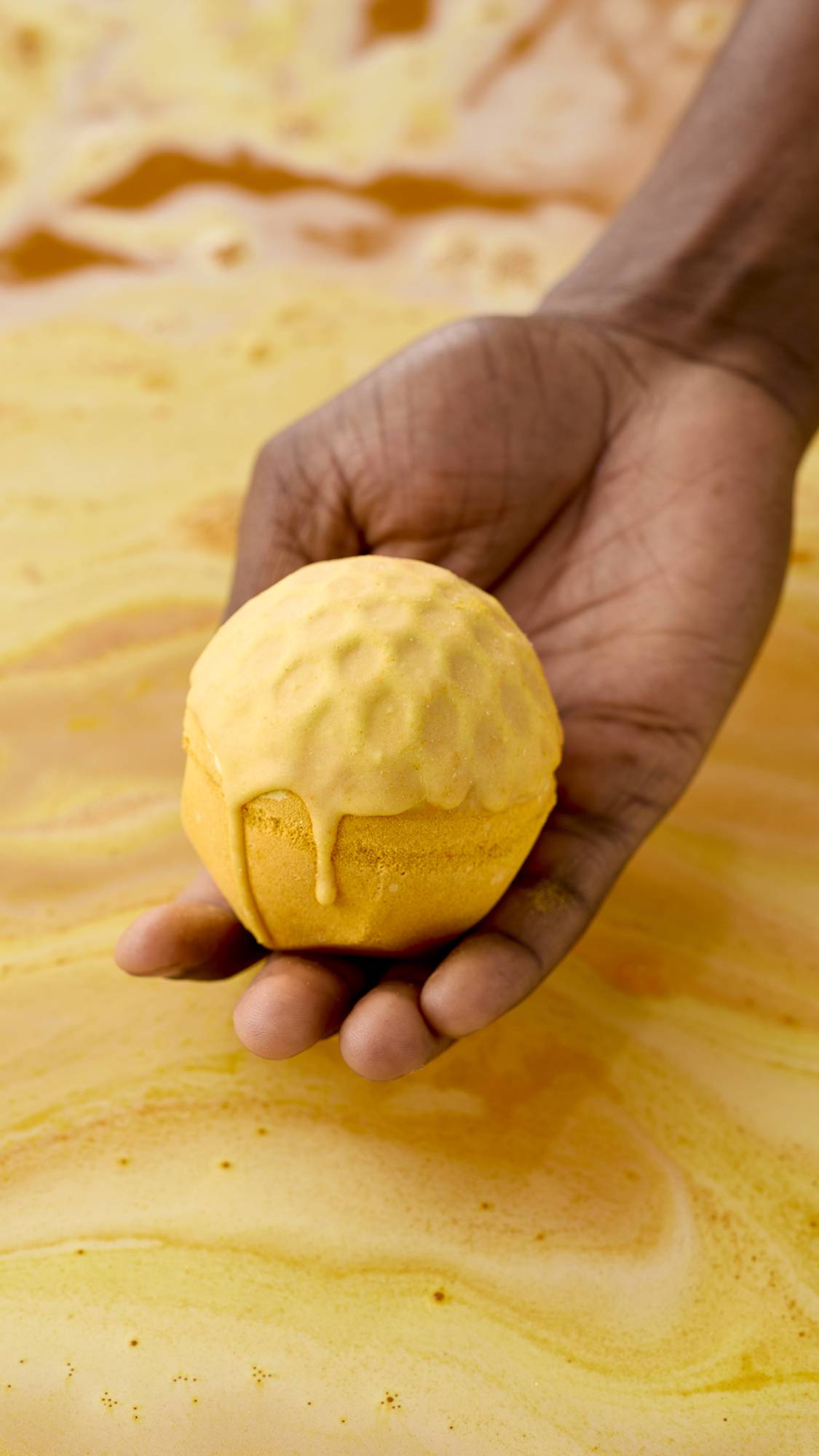 A hand holds a golden-yellow, "Sweet As Honey" bath bomb over a tub of swirling, amber-coloured water. The top of the bath bomb appears to be melting or dripping down the sides, set against the vibrant orange bathwater background.