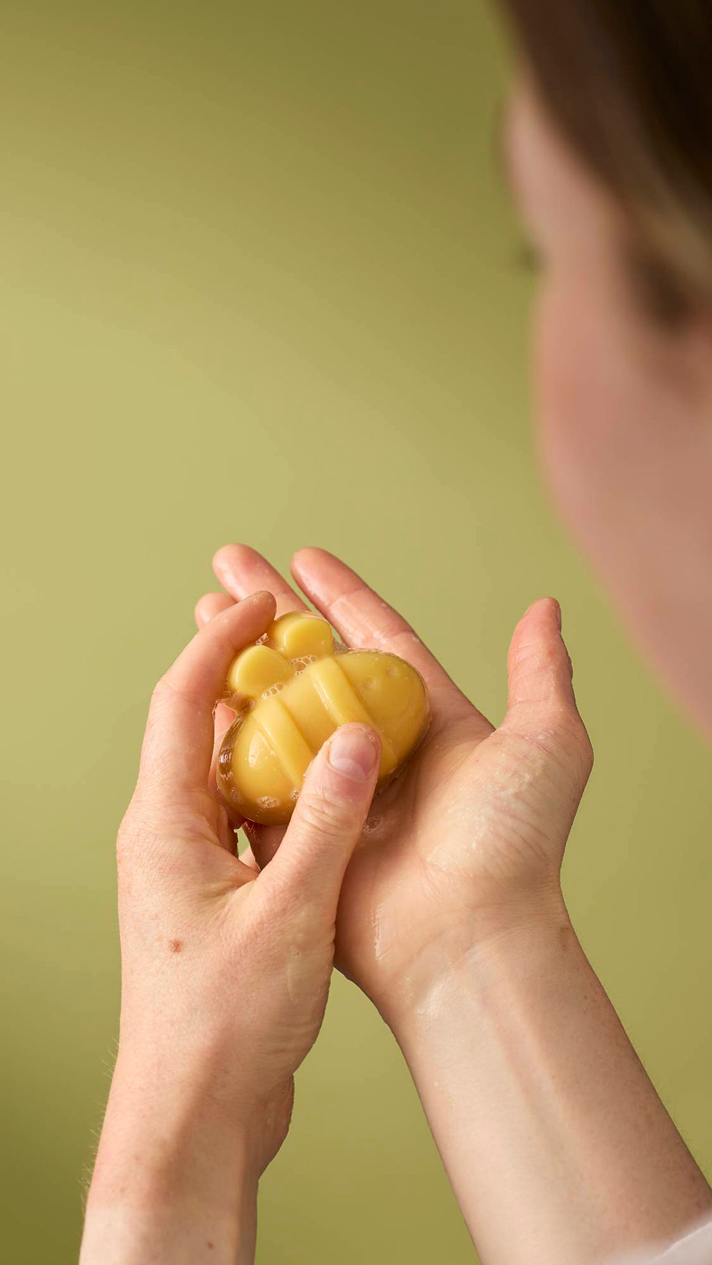 A person holds a small, yellow, bee-shaped facial gummy wash in their wet hands. The product is slightly bubbly, and the person appears to be lathering it against an olive-green background.