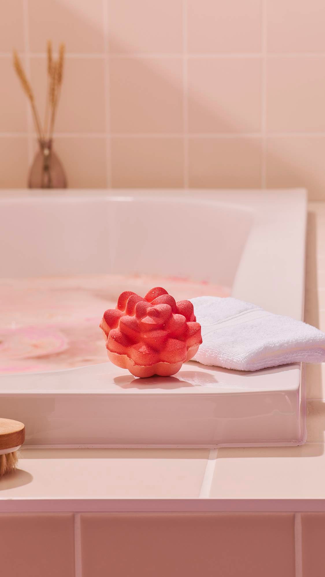A vibrant, pink bath bomb shaped as a blooming flower sits on the corner of a white bathtub. A folded white flannel lies next to it, with the soft pink water of a bath visible in the background.
