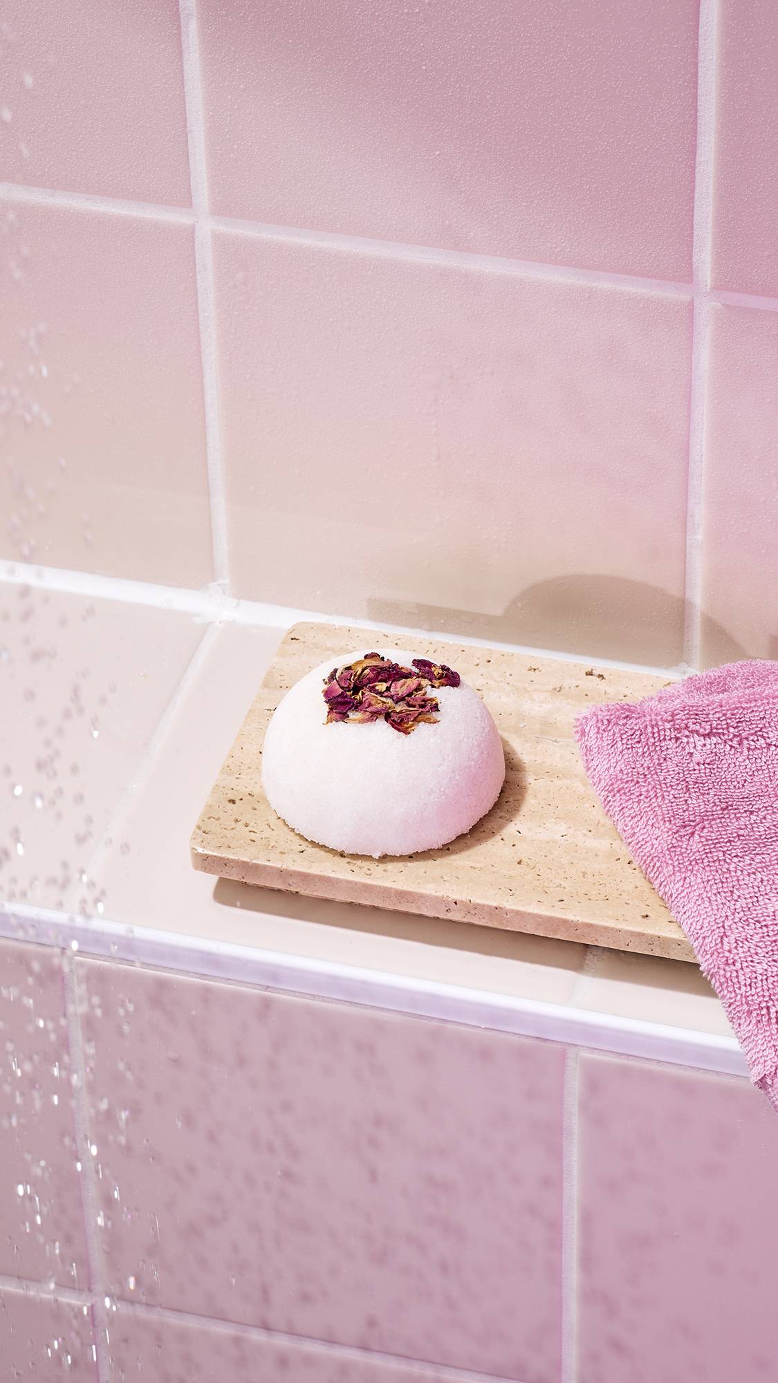 The image shows the "Comforter Rose" solid body scrub topped with dried rose petals, sitting on a beige stone tray in a bathroom. A pink flannel is tucked beside it, set against a background of soft pink wall tiles.