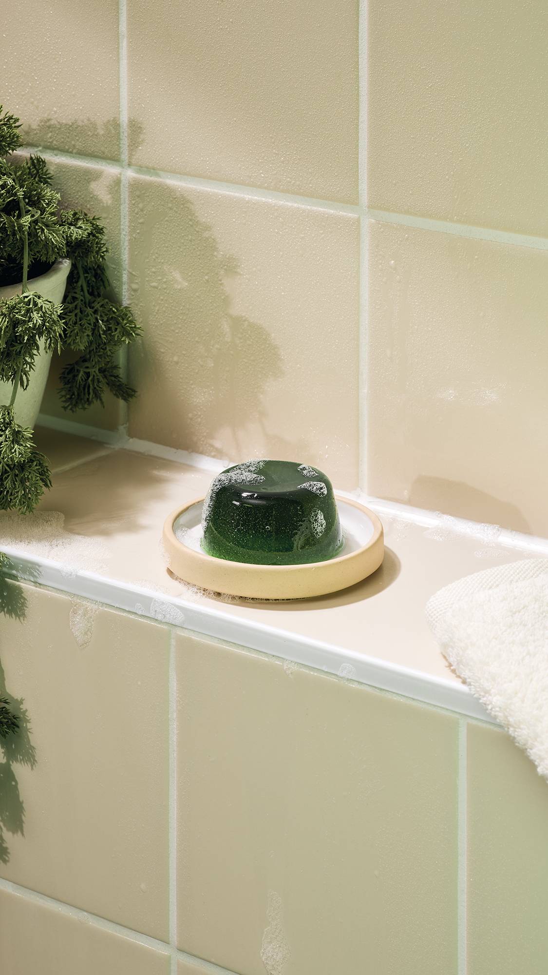 The image shows the green, translucent "The Good Hour" shower jelly sitting on a ceramic dish on a bathroom ledge. The jelly has a smooth, wobbly dome shape with bubbles on its surface, set against a background of neutral-toned wall tiles.