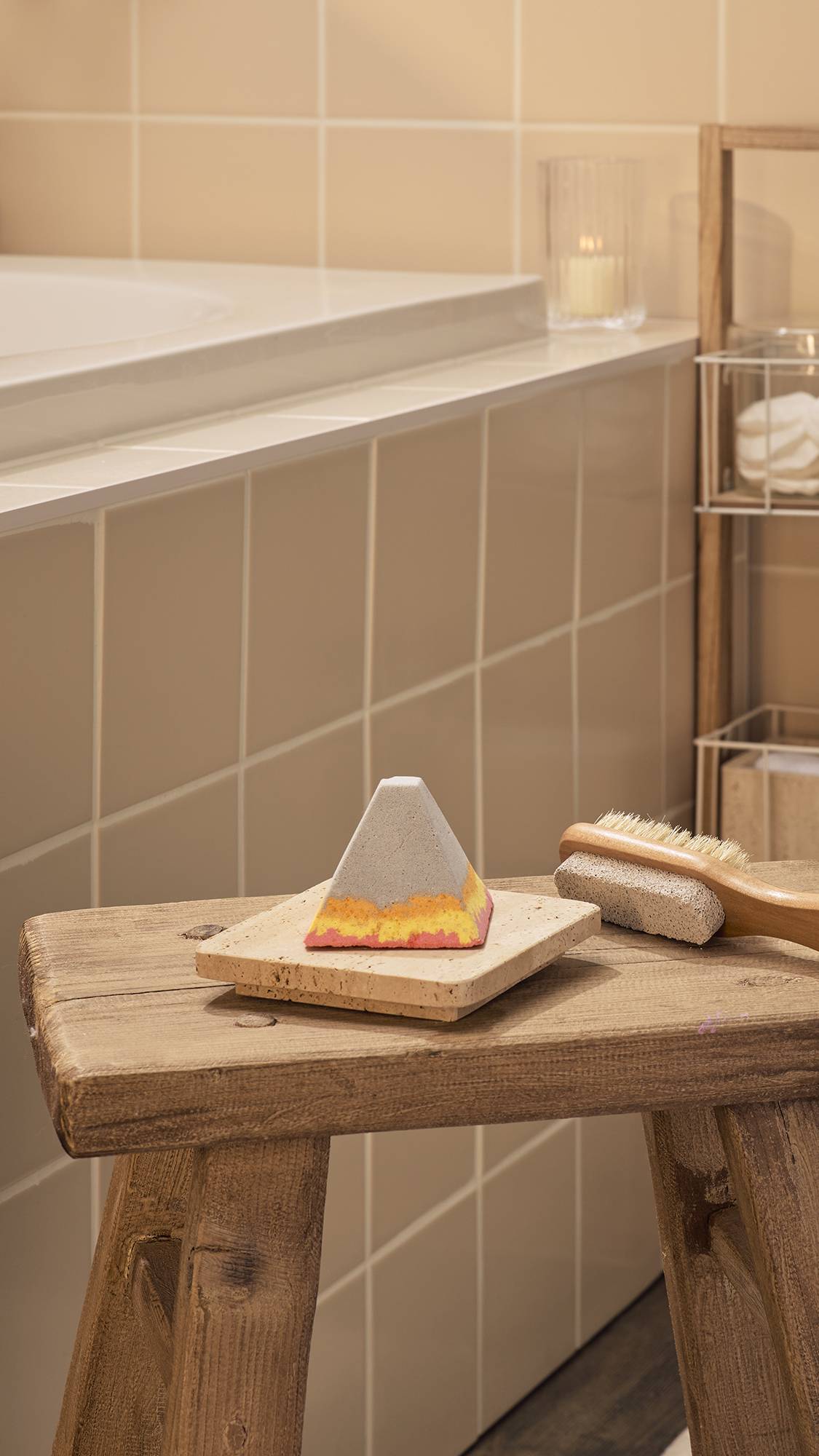The "Volcano", a pyramid-shaped bath bomb-style foot soak with a grey top and wavy layers of orange, yellow, and pink at the base, sits on a stone tray positioned on a rustic wooden stool in a bathroom with neutral wall tiles and a lit candle.