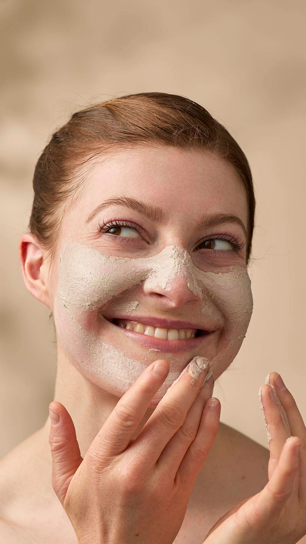 A close-up of a smiling model with reddish-brown hair tied back, applying a textured, beige face mask. They look off-camera with a playful expression while using their fingers to blend the thick cleanser across their nose, cheeks, and chin.