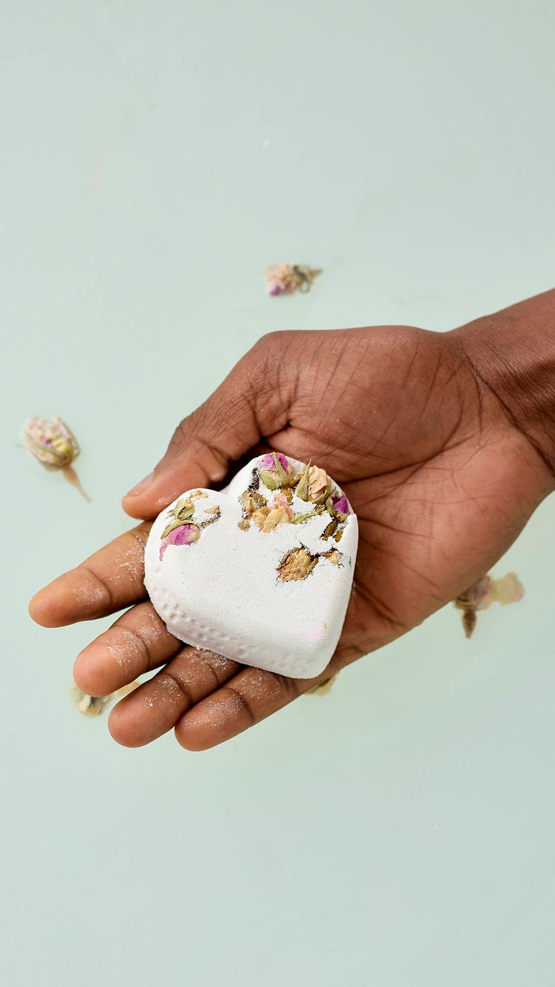 A hand holds a white, heart-shaped Tisty Tosty bath bomb decorated with small dried rosebuds and petals. Several loose rosebuds are scattered on the pale water surface in the background.