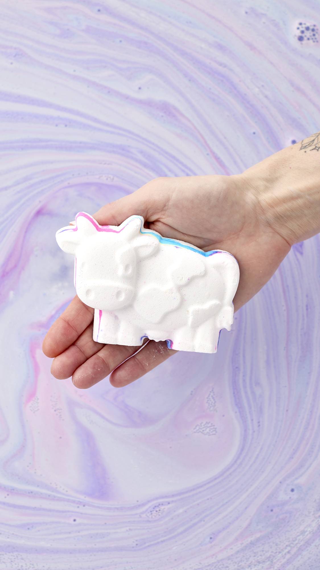 A hand holds the white, cow-shaped Toby's Magic Cow Bath Bomb over a bathtub filled with swirls of pastel purple and blue water. This bath bomb features pops of hidden colors along its edges, ready to release a rainbow of foam into the tub.