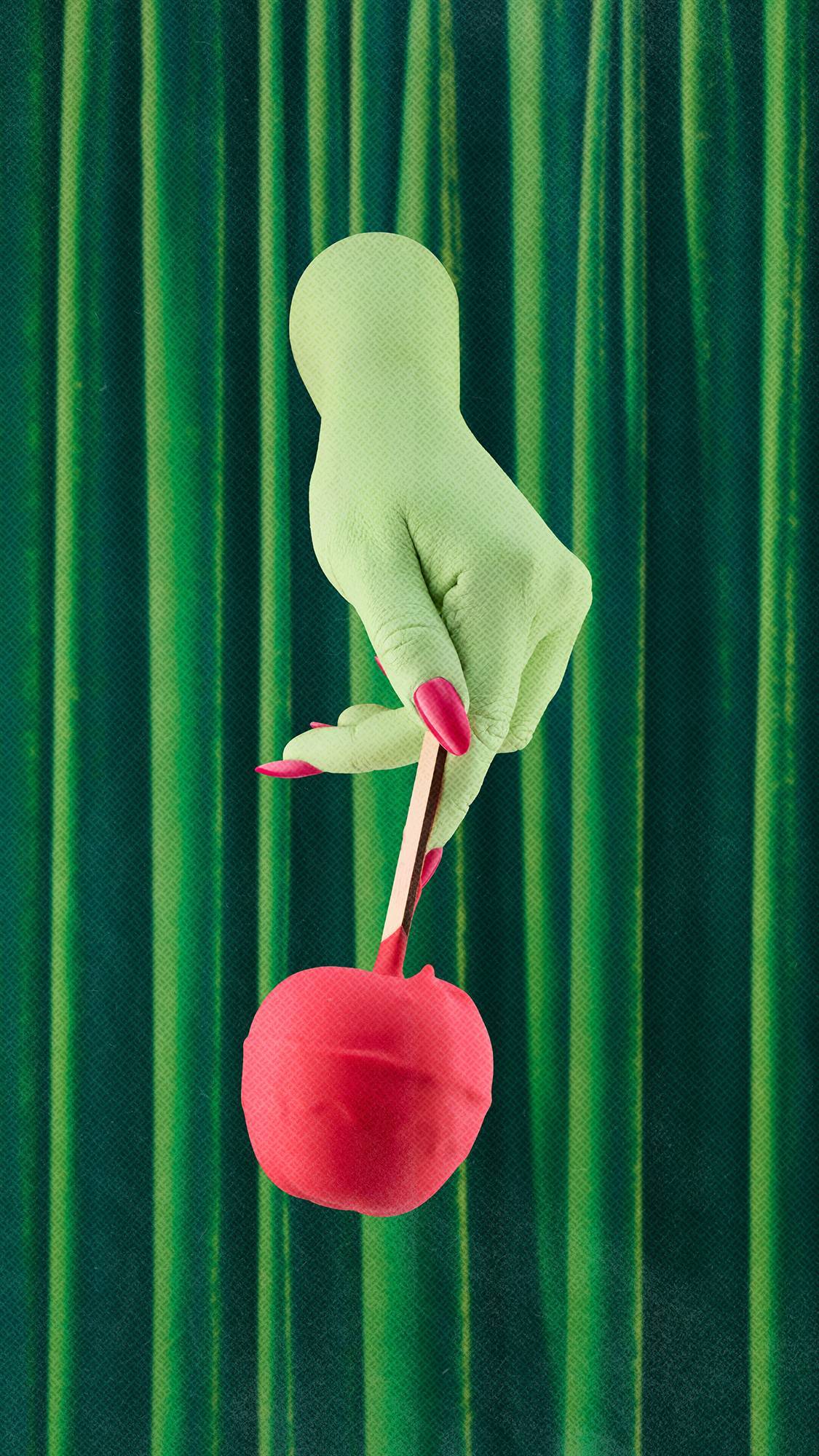 A pale green hand with bright pink fingernails holds a red toffee apple bath bomb on a wooden stick, pointing downwards. The hand is positioned against a background of draped green velvet curtains.