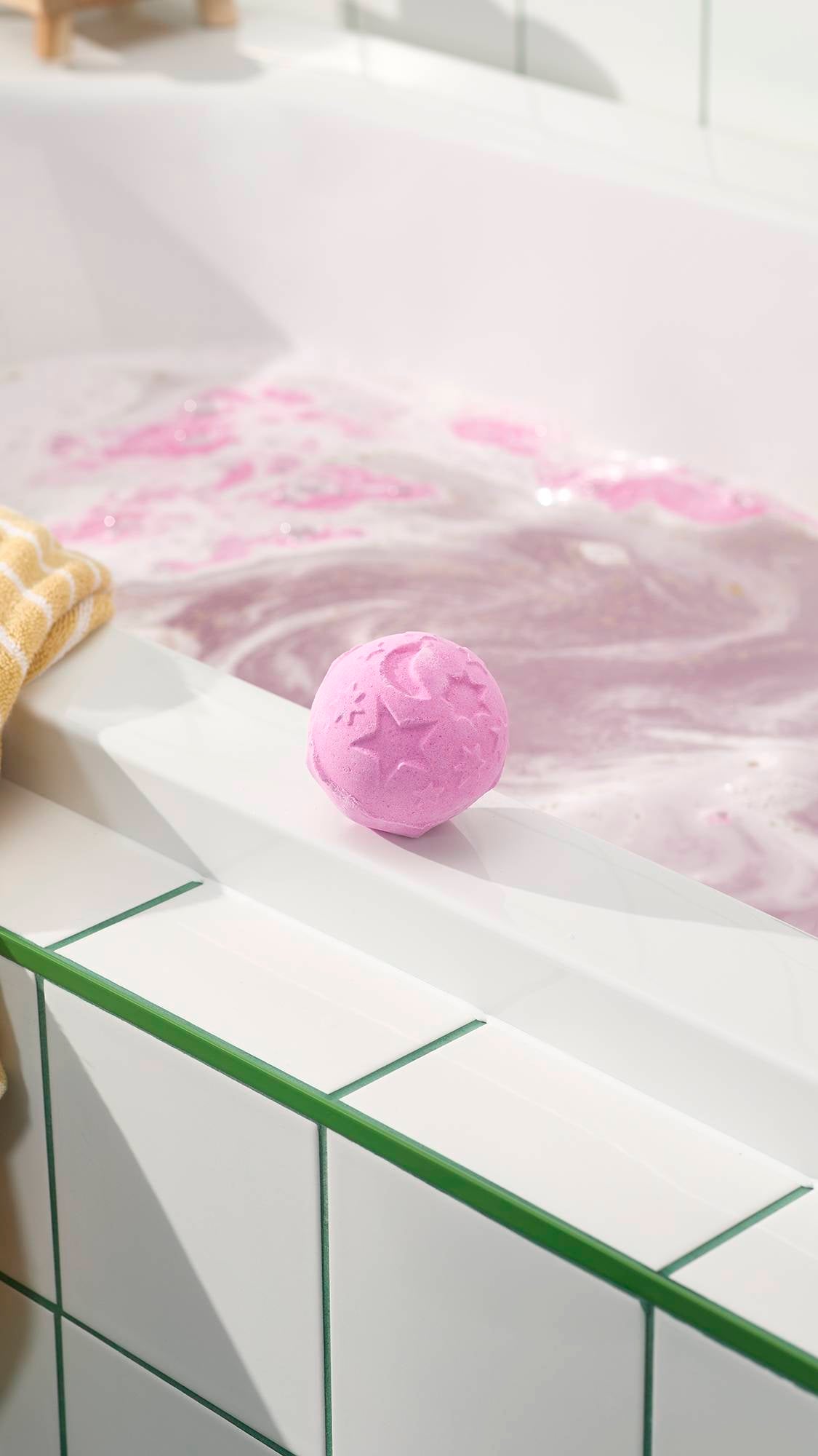 The Twilight Bath Bomb sits on the clean, white edge of a bathtub, overlooking water already filled with swirling lavender and pink foam. This celestial-themed bath bomb is a soft purple sphere decorated with embossed stars and a crescent moon.