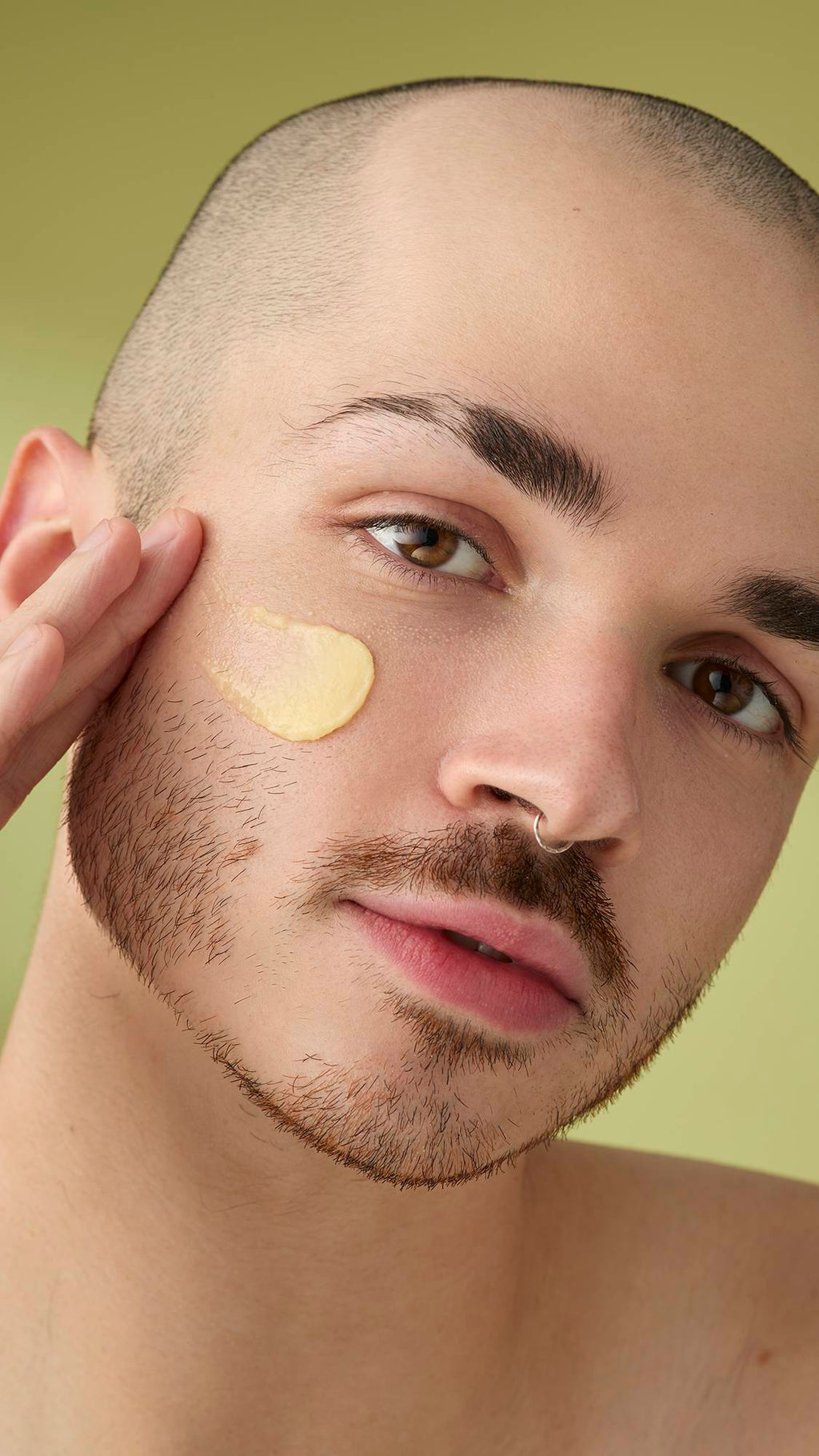 A close-up portrait of a model with a shaved head and facial hair, who has a smear of pale yellow 'Ultraplant' facial cleanser on their cheek. They are looking into the camera with a neutral expression against a soft olive-green background.