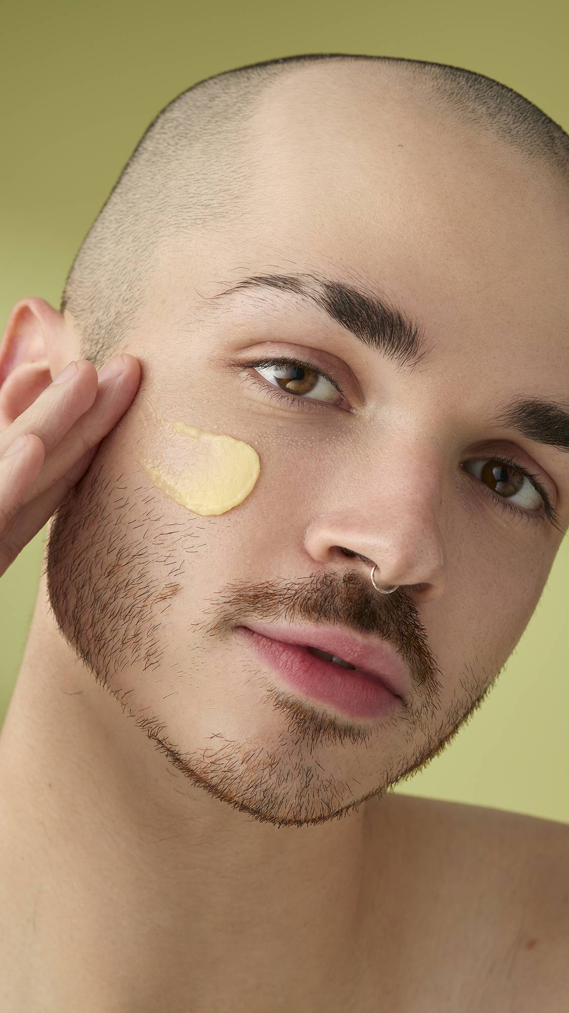 A close-up portrait of a model with a shaved head and facial hair, who has a smear of pale yellow 'Ultraplant' facial cleanser on their cheek. They are looking into the camera with a neutral expression against a soft olive-green background.