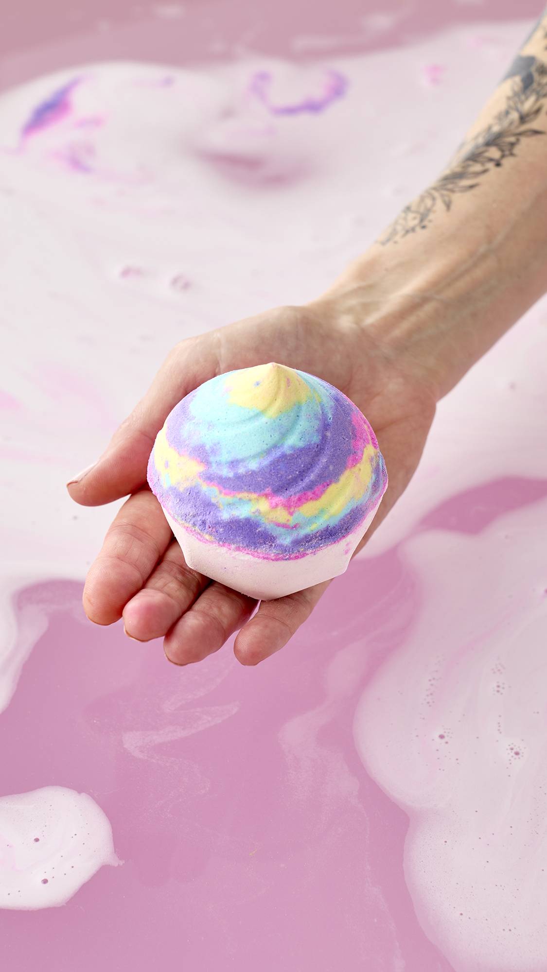 A hand holds the Unicorn Poop Bath Bomb over a bathtub filled with gently swirling pink and purple water. The whimsical, swirl-shaped bath bomb features a white base topped with a vibrant mix of pastel blue, yellow, purple, and pink hues.
