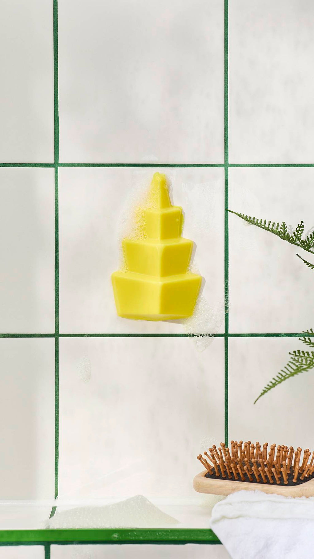 A tiered, bright yellow geometric shower gummy soap stuck to white bathroom tiles with green grout, surrounded by soapy bubbles. A wooden hairbrush and fern are visible below.