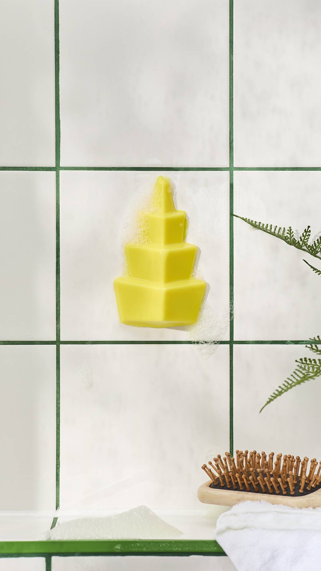 A tiered, bright yellow geometric shower gummy soap stuck to white bathroom tiles with green grout, surrounded by soapy bubbles. A wooden hairbrush and fern are visible below.