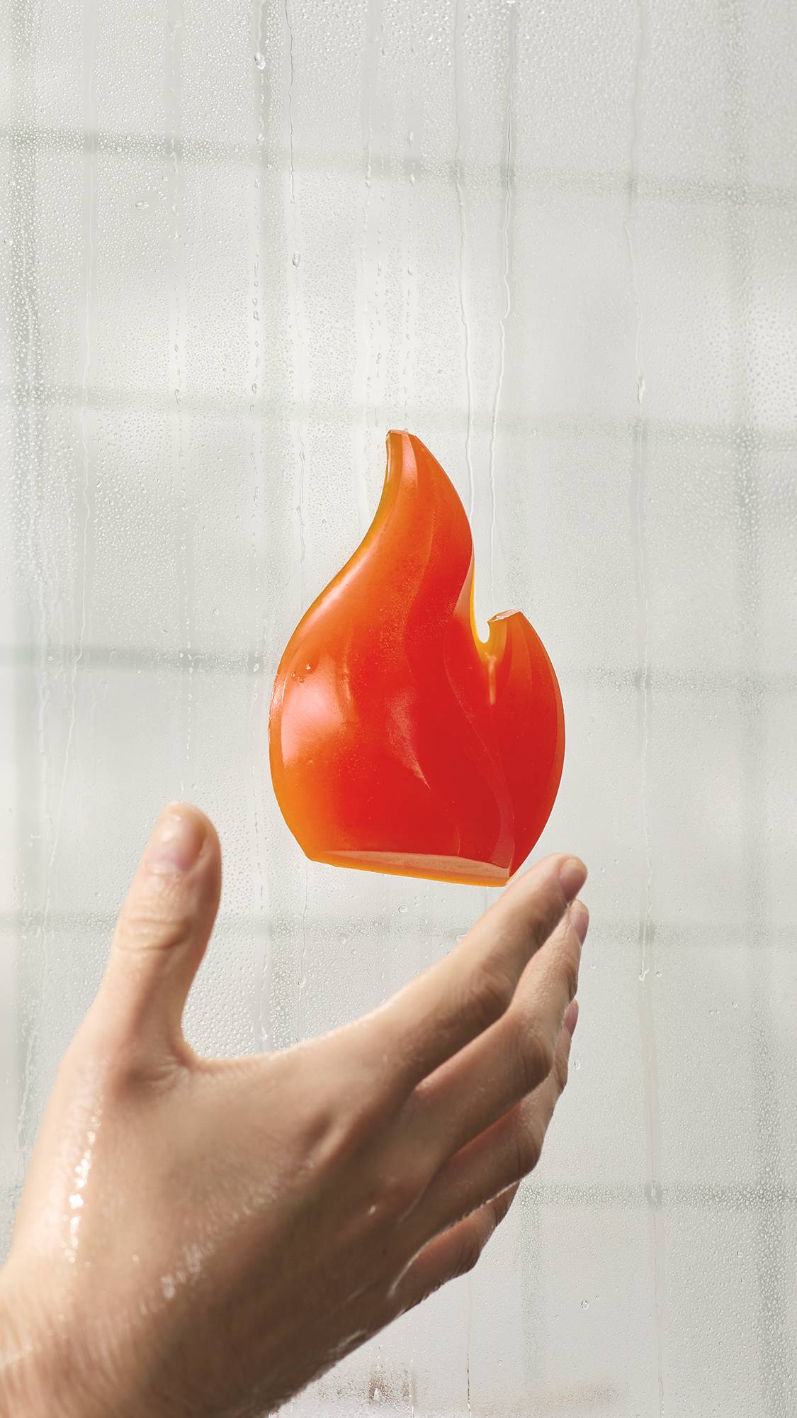 A bright orange, flame-shaped shower gummy soap stuck to a steamy glass shower door. A wet hand reaches toward it from below against a blurry background of white bathroom tiles.