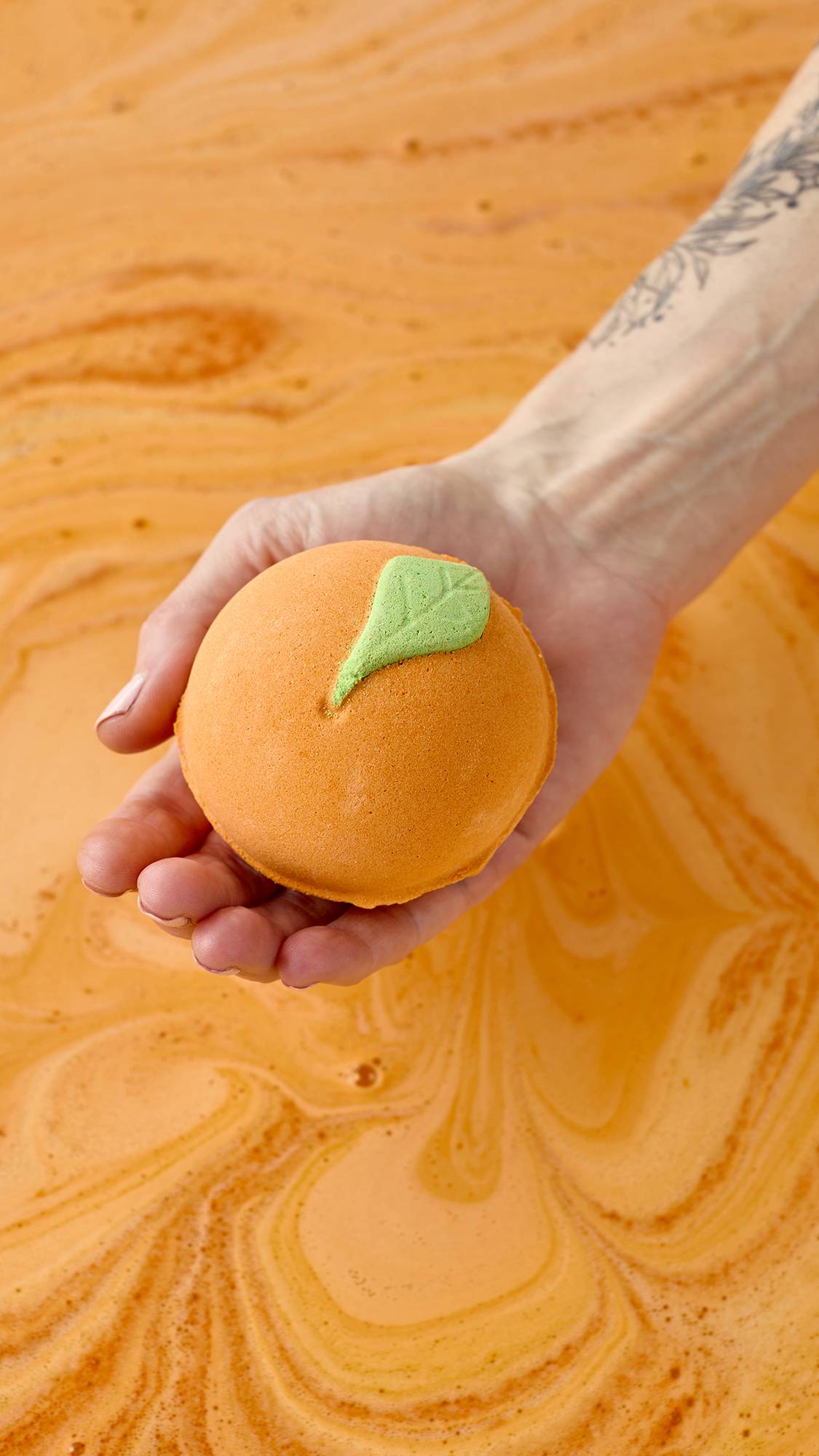 A hand holds the Peachy Bath Bomb over a tub of swirling orange water. The orange sphere, shaped like a real peach with a green leaf, creates a vibrant, citrusy soak.