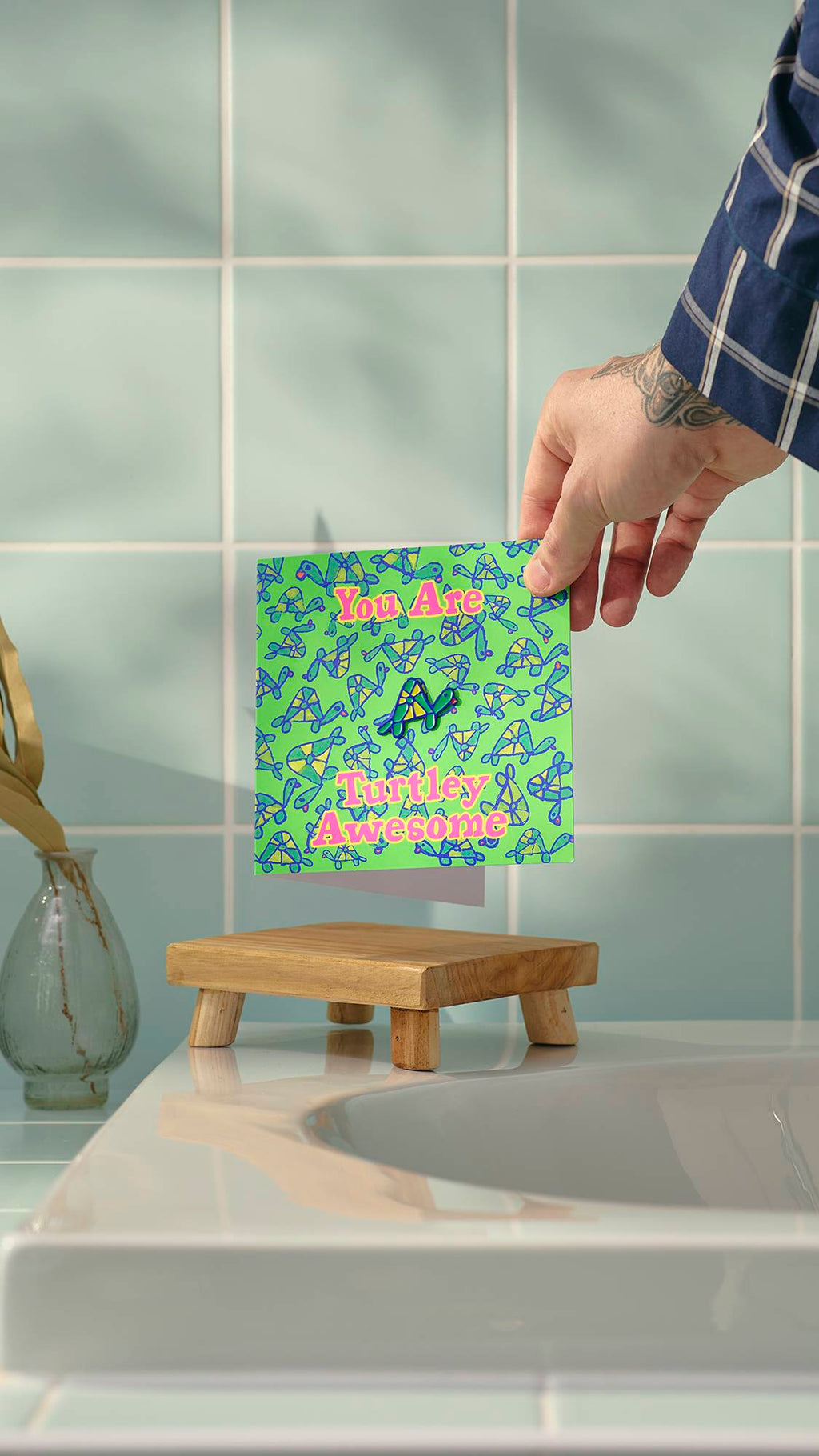 A tattooed hand holds a ‘Turtley Awesome’ greeting card over a wooden soap dish in a bathroom. The green card features a small turtle enamel pin and a repeating turtle pattern, set against a background of pale blue wall tiles.