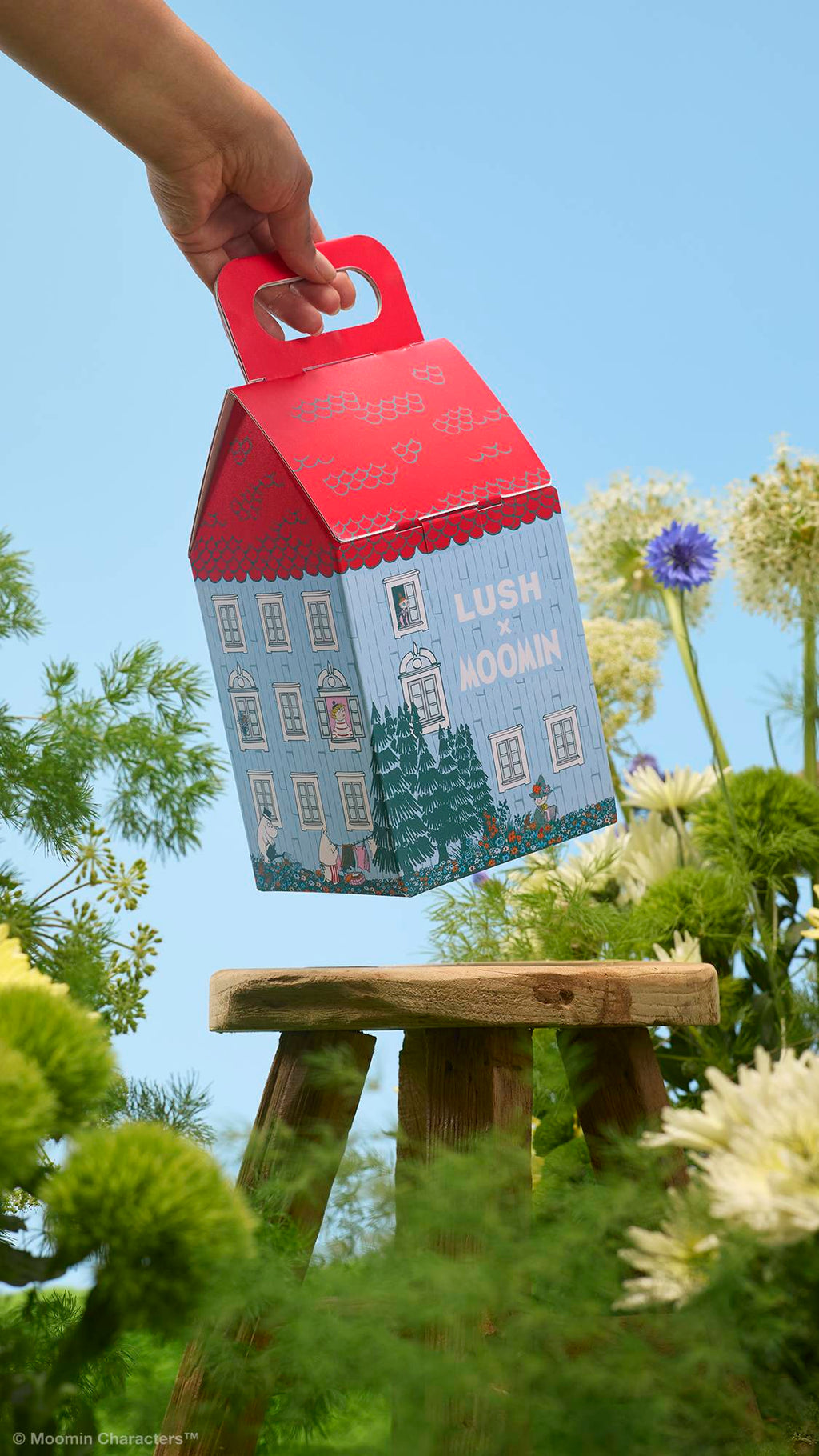 A hand holds a house-shaped "LUSH x MOOMIN" gift box by its red handle above a small wooden stool. The background features lush green foliage and wildflowers against a clear blue sky.