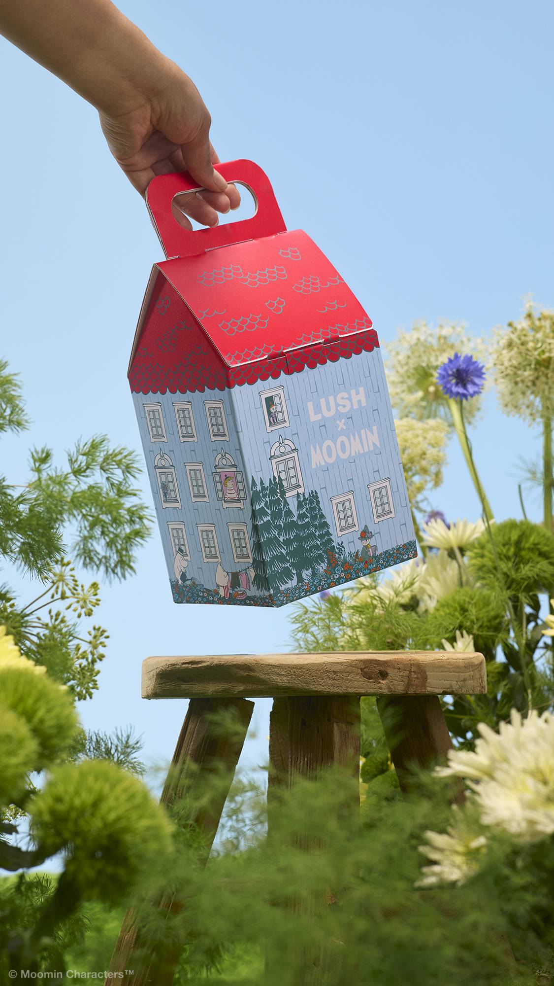 A hand holds a house-shaped "LUSH x MOOMIN" gift box by its red handle above a small wooden stool. The background features lush green foliage and wildflowers against a clear blue sky.