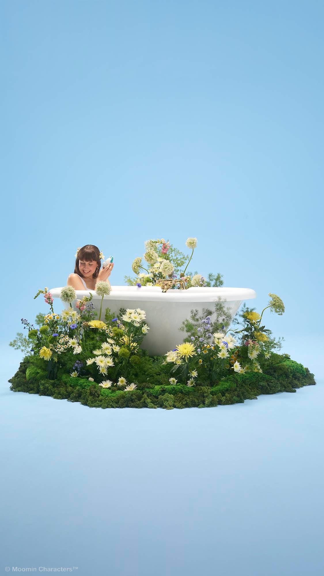 A person smiles whilst sitting inside a white bathtub, holding a small bath bomb. The tub is placed on a patch of lush green moss overflowing with yellow and white wildflowers, set against a plain light blue background.