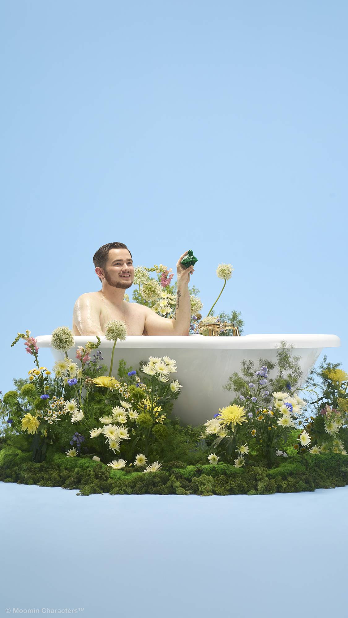 A person smiles whilst sitting inside a white bathtub, holding up a small green Snufkin-shaped shower gummy. The tub sits on a patch of lush green moss filled with yellow and white wildflowers, set against a plain light blue background.