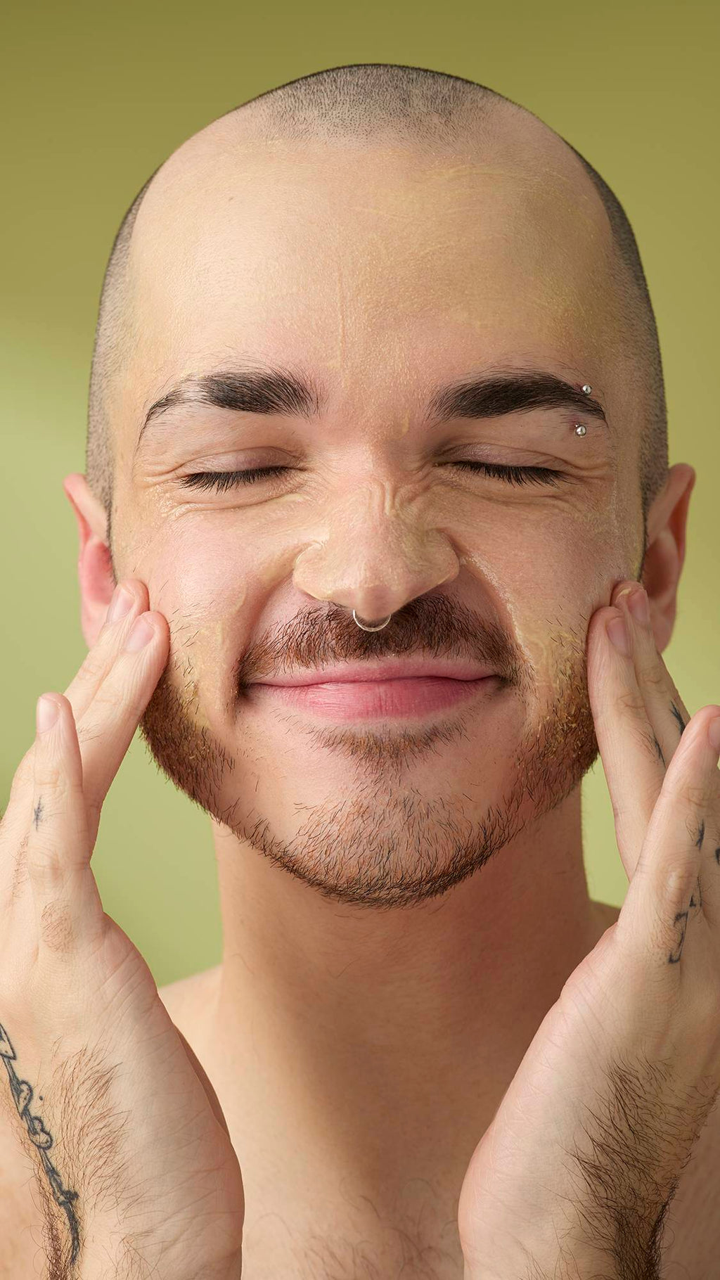 A person with a shaved head and facial piercings smiles with their eyes closed while massaging the Acai And Sacha Inchi cleanser into their face. They have tattooed hands and are shown from the chest up against a plain olive-green background.