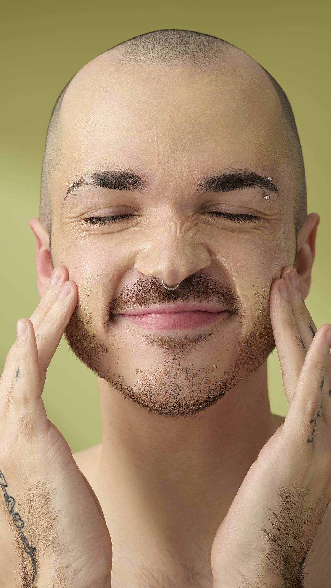 A person with a shaved head and facial piercings smiles with their eyes closed while massaging the Acai And Sacha Inchi cleanser into their face. They have tattooed hands and are shown from the chest up against a plain olive-green background.