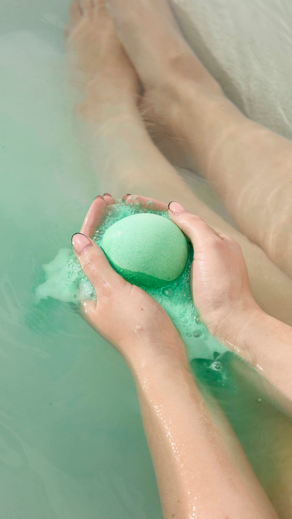 Hands hold the bright green Avobath Bath bomb as it fizzes in a tub of pale green water. A stream of frothy, light green bubbles flows from the bomb, with submerged legs visible in the background.