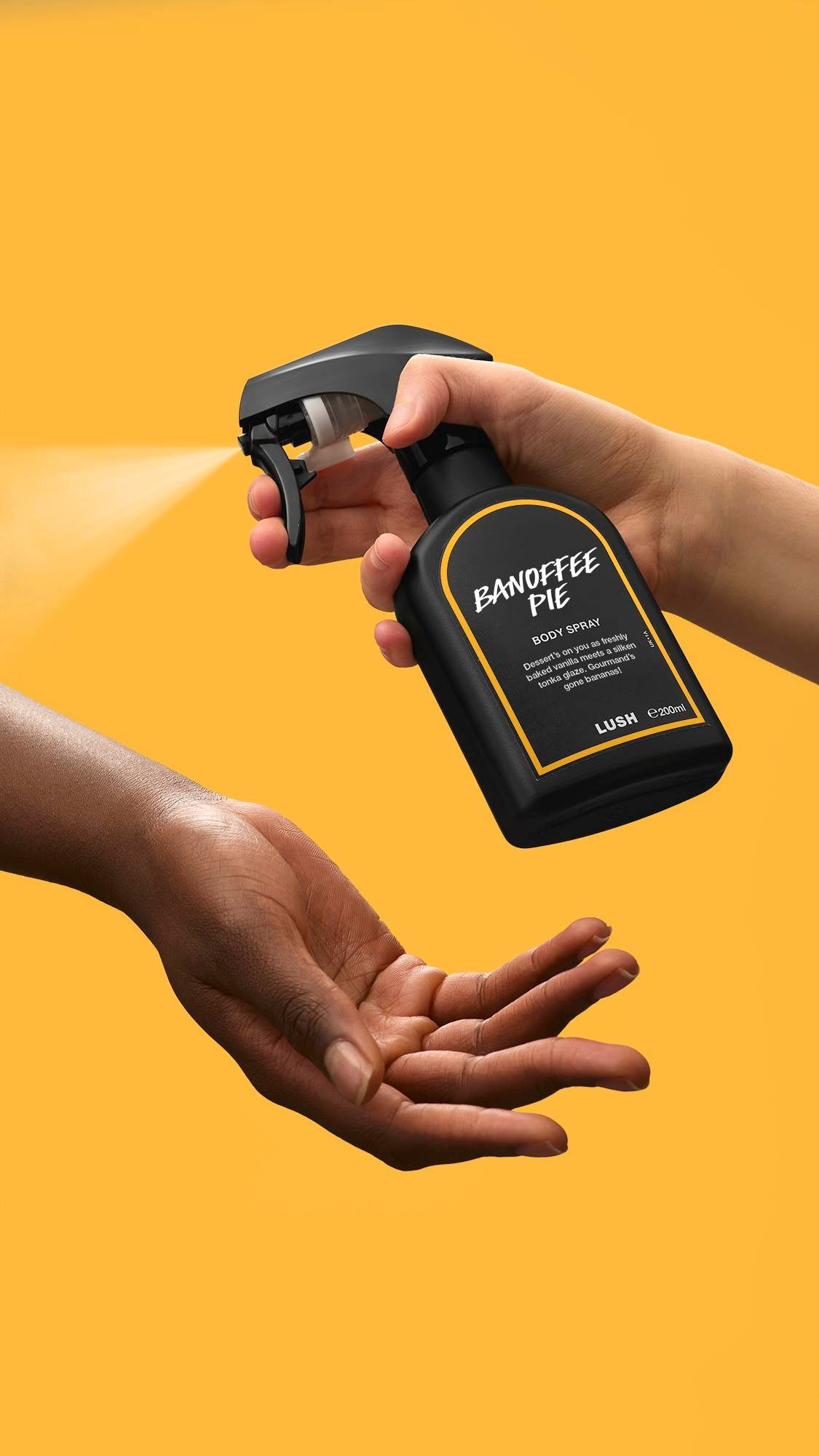 A person's hand presses the nozzle of a black Lush 'Banoffee Pie' body spray bottle, releasing a fine mist onto another person's open palm. The action is set against a solid, vibrant yellow background.