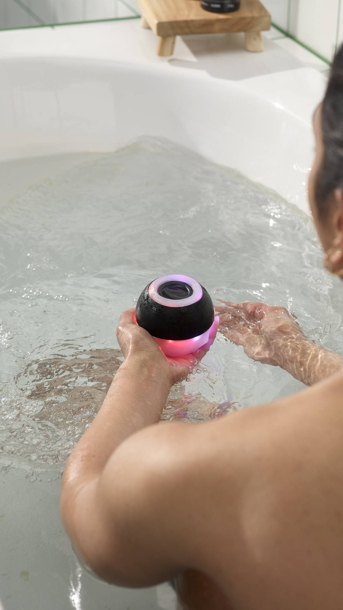 A model in a bathtub holds a black, spherical, bath bomb-shaped waterproof speaker that glows with pink and purple light. The device is partially submerged in the clear bathwater, with ripples expanding around the person's hands.