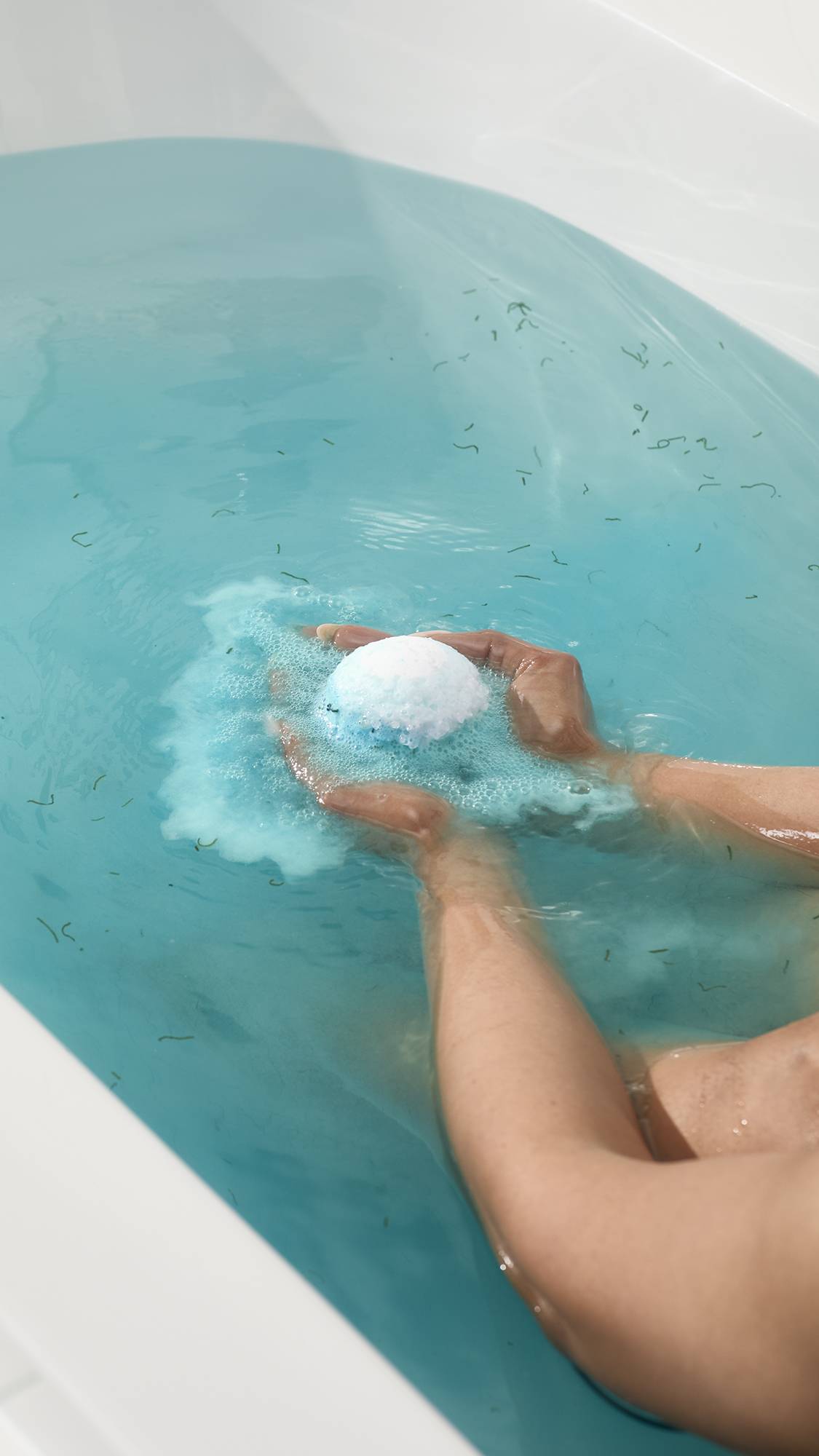 Hands hold the Big Blue bath bomb as it fizzes in a tub of bright blue water. A cloud of frothy blue foam surrounds the bomb, while small green pieces of seaweed float throughout the water.