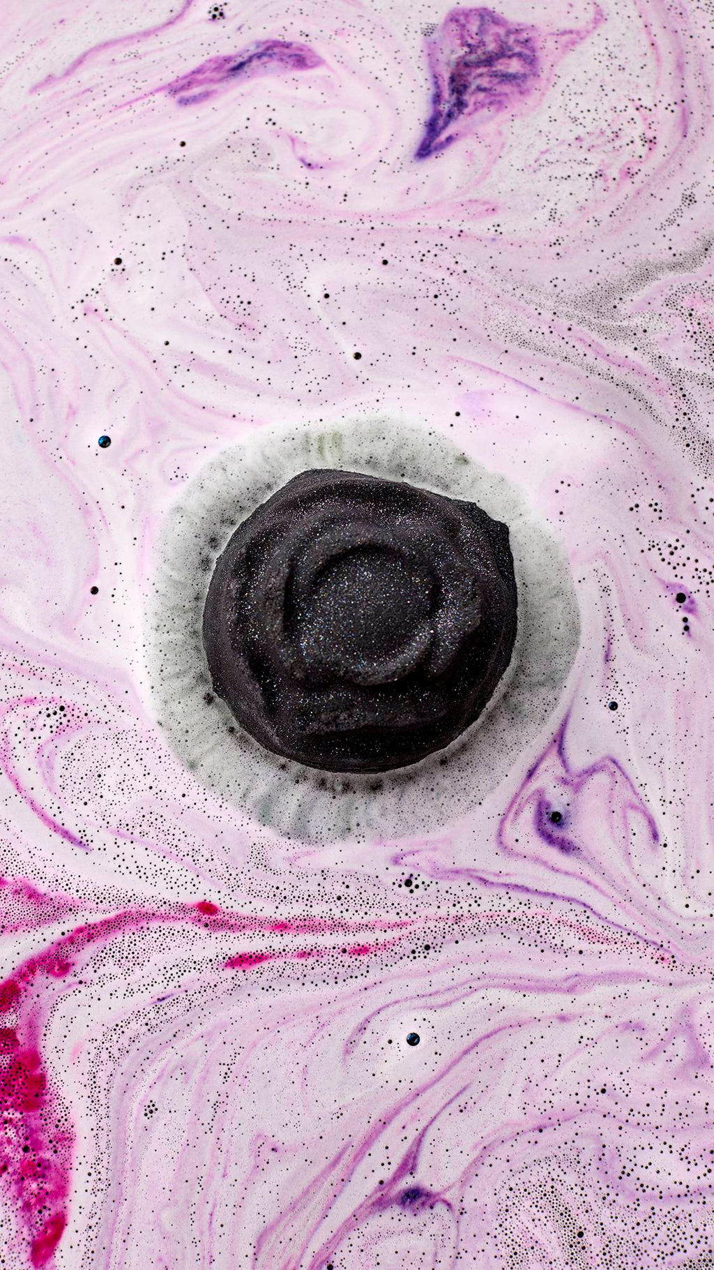 Black Rose. An aerial view shows the dark rose-shaped bath bomb as it dissolves. It creates a thick layer of velvety, deep-purple foam on the water's surface, rippling with bright pink swirls and shimmering silver plastic-free glitter.