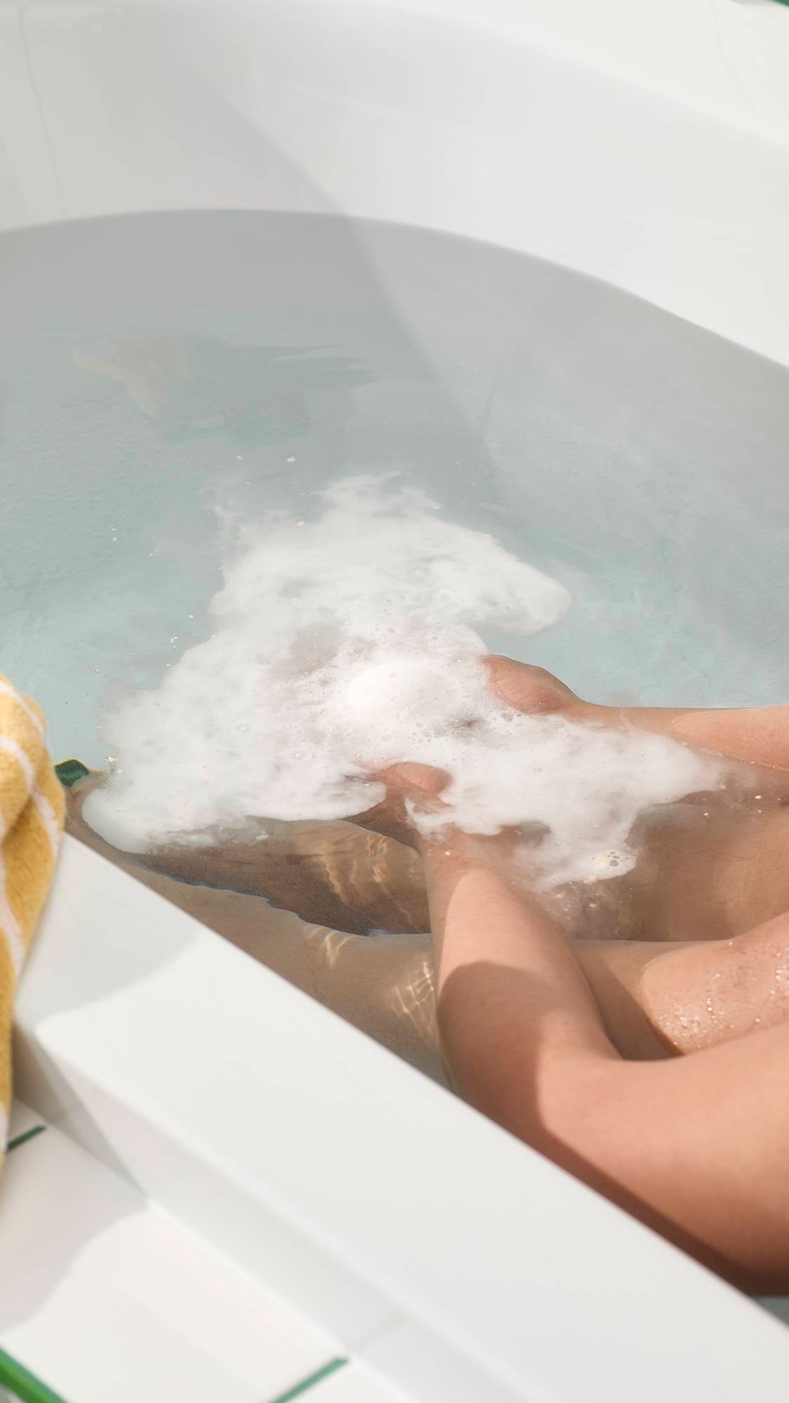 An aerial view of the white, spherical Butterball bath bomb dissolving in light blue water. The product creates a layer of creamy, white foam on the surface, with visible small pieces of cocoa butter floating as it fizzes.