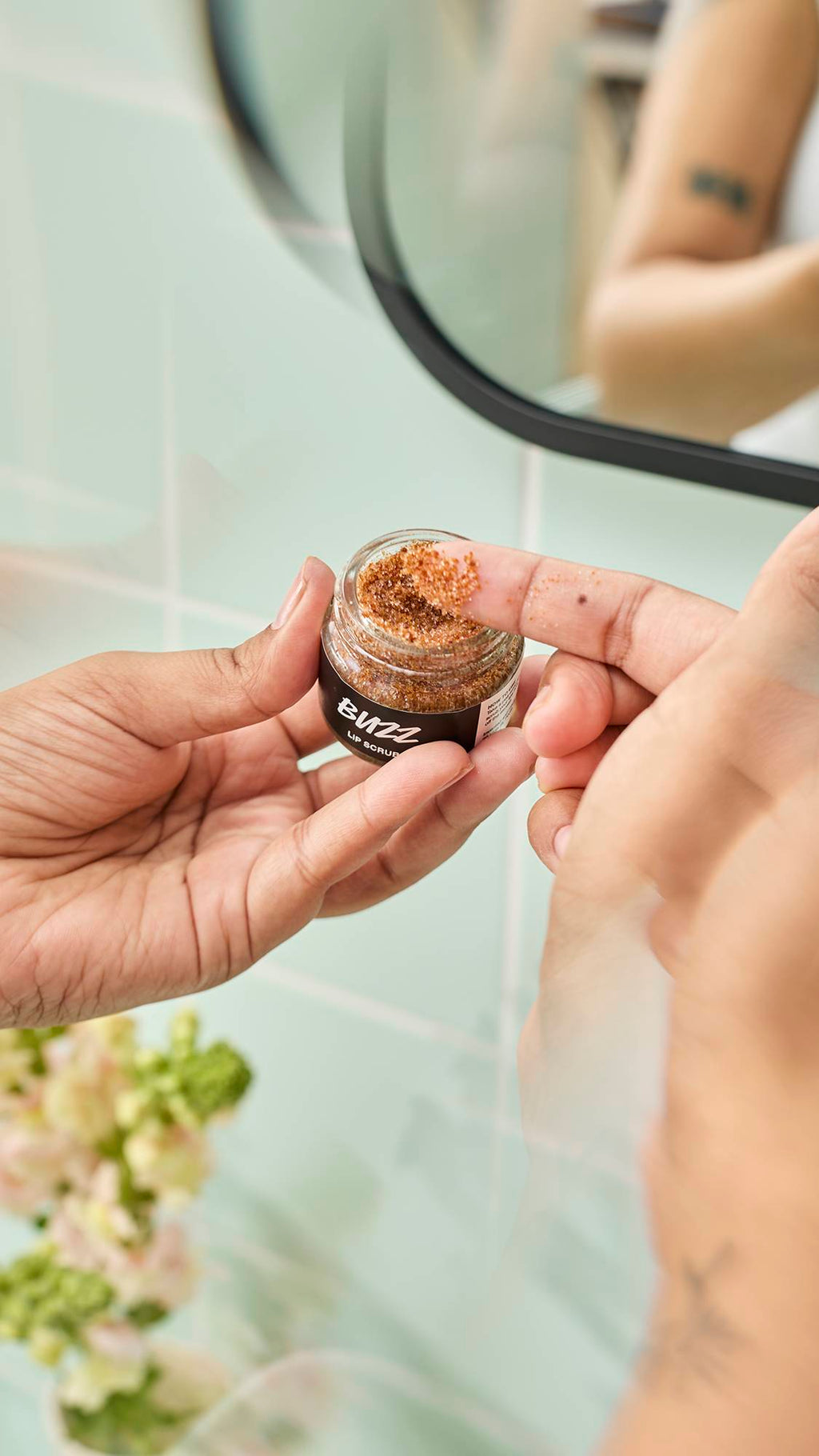 A close-up shot shows a model's finger dipping into a small glass jar of Lush "Buzz" Lip Scrub to scoop out the textured, caramel-coloured product. The scene is set against a background of pale green bathroom tiles and soft floral decorations.