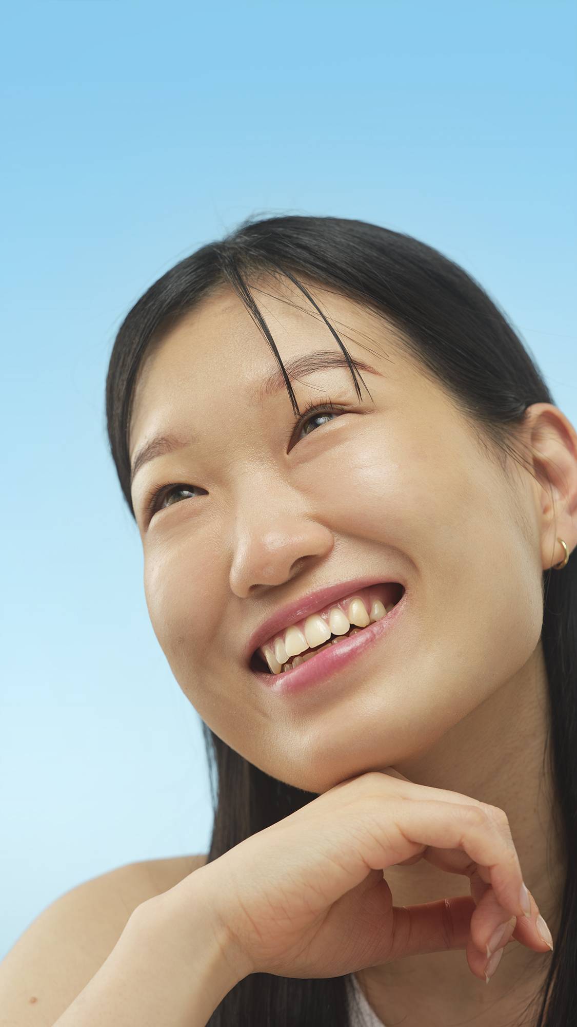 A close-up vertical portrait of a smiling person with dark hair, looking up and left. Their chin rests on their hand, and their face has a healthy, radiant sheen from the application of the Calm primer, set against a bright blue background.