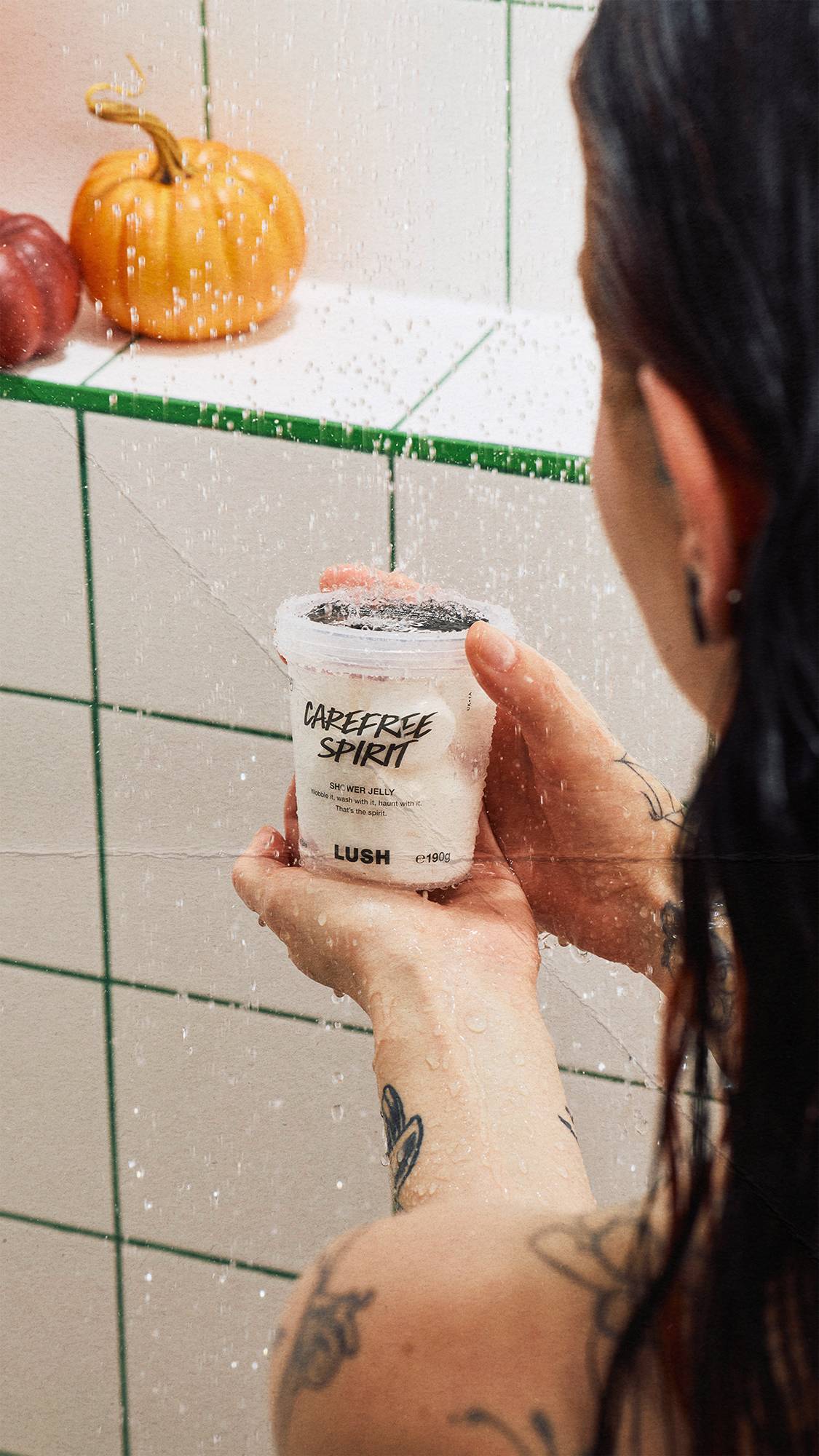 A person with tattoos holds a clear pot of Lush 'CAREFREE SPIRIT' shower jelly under running water in a shower. White tiles with green grout form the background, with decorative pumpkins on a ledge behind them.
