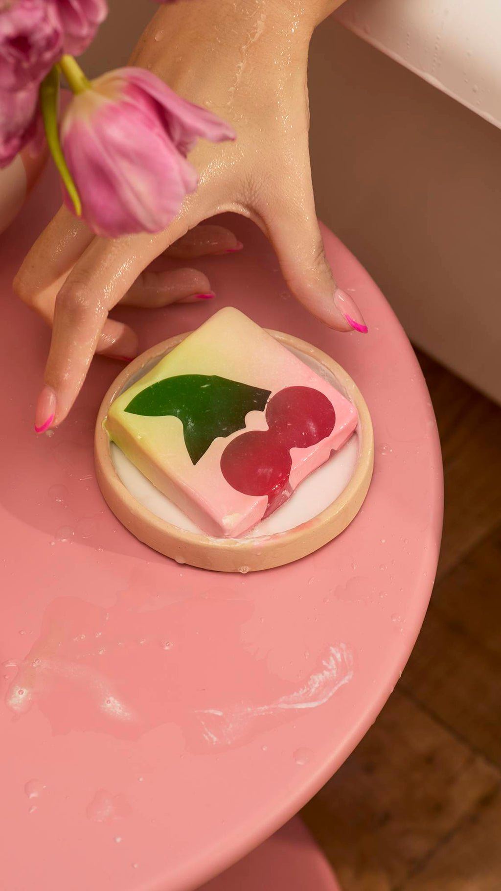 A square Cherry On Top soap bar with a dark cherry and green leaf design sits in a small dish on a pink table. A person with pink-tipped nails reaches for the soap near a white bathtub and a vase of pink flowers.
