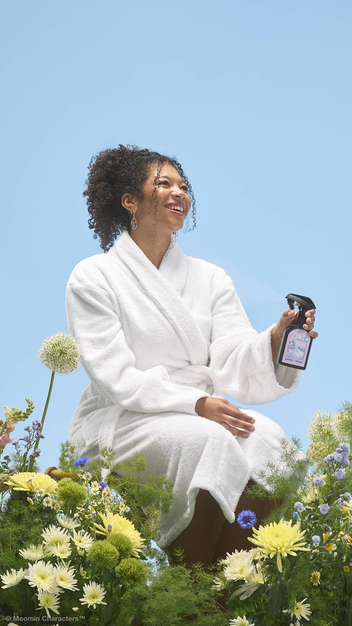 A close-up shot of a smiling person wearing a white dressing gown, sitting amidst a vibrant patch of wildflowers. They hold a black bottle of "Cloudberry" body spray, spritzing a light mist into the air against a clear blue sky background.