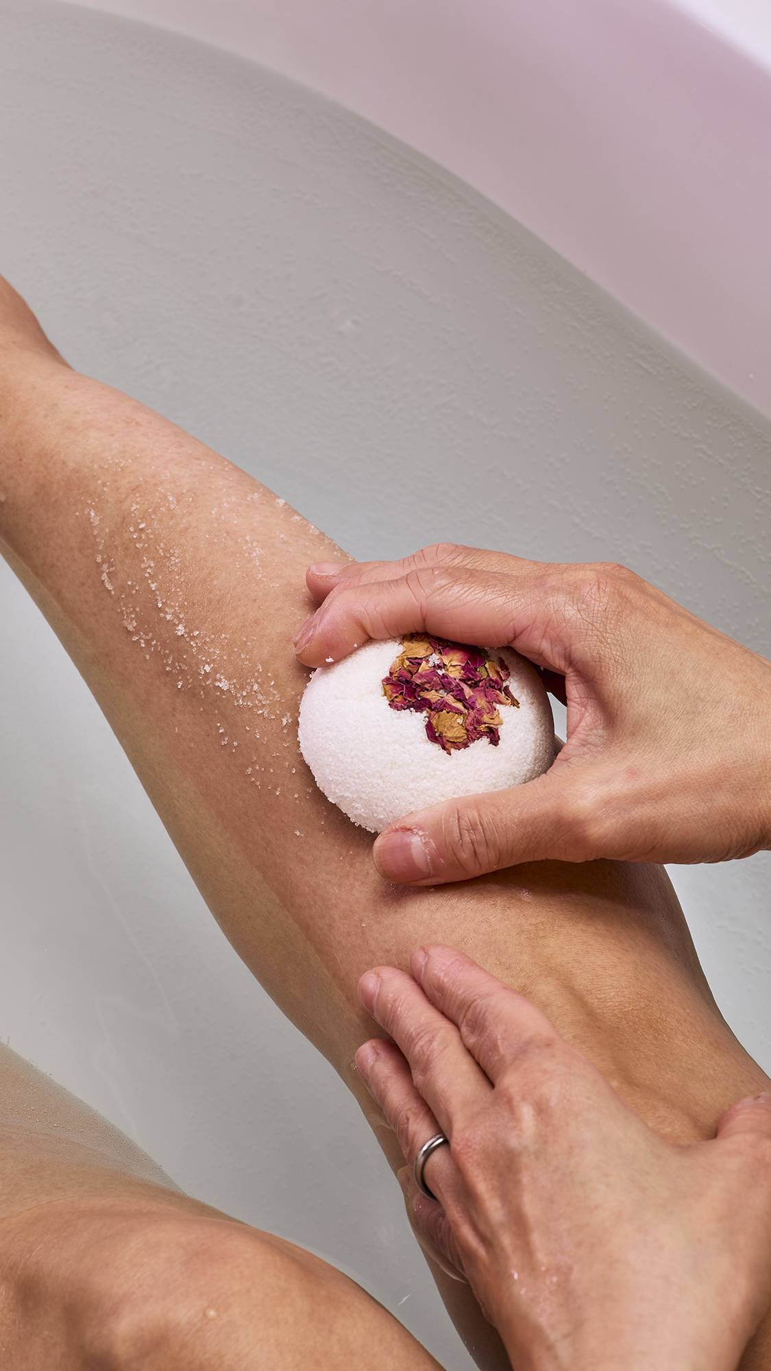 A model in a bath uses the white, dome-shaped "Comforter Rose" solid body scrub. They are rubbing the textured surface against their leg, leaving a fine, white exfoliating residue on the skin.