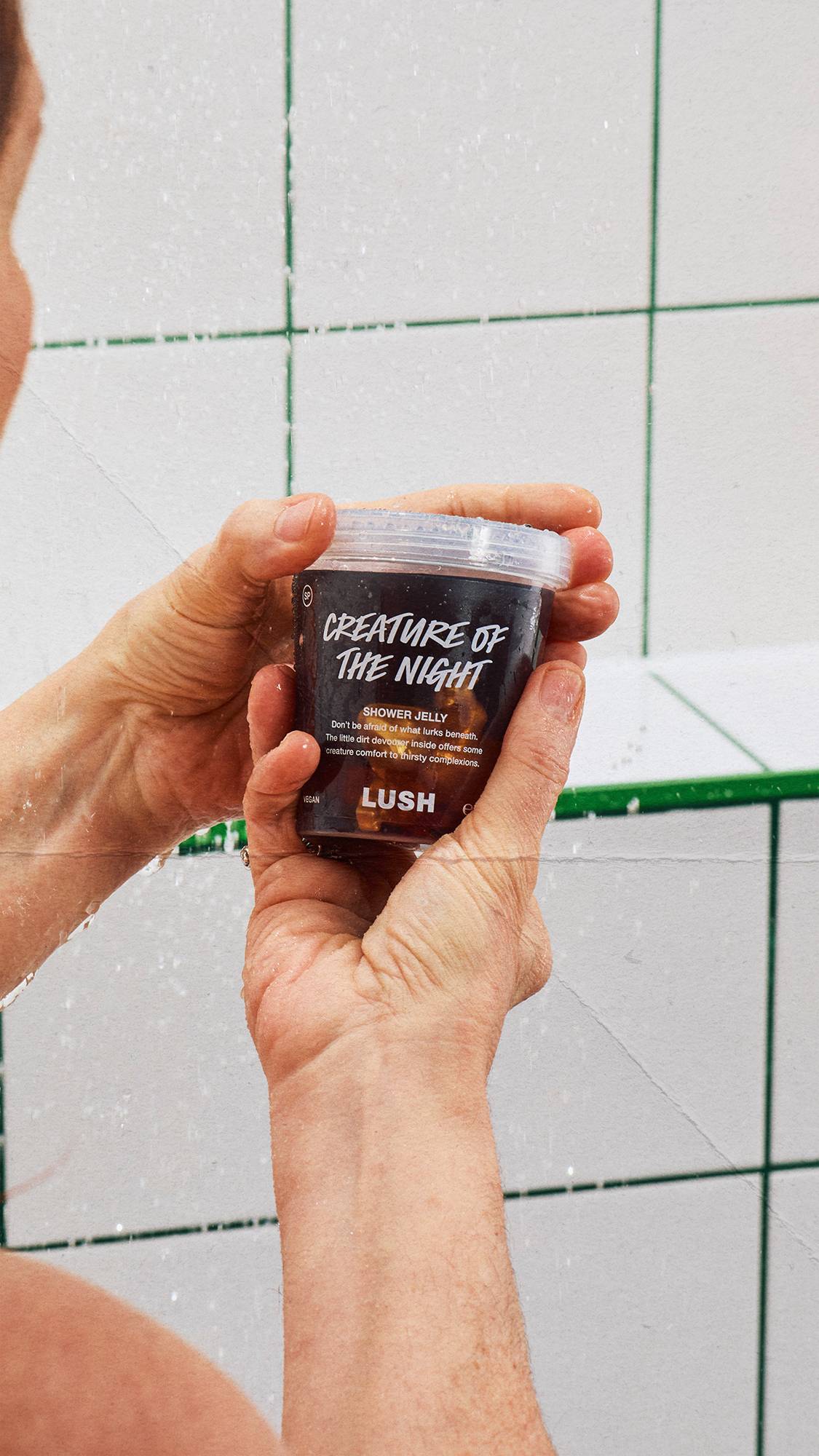 Two hands hold a clear tub of Lush 'CREATURE OF THE NIGHT' shower jelly under a running shower. Water drops splash across the container. The background shows white square tiles with green grout lines.