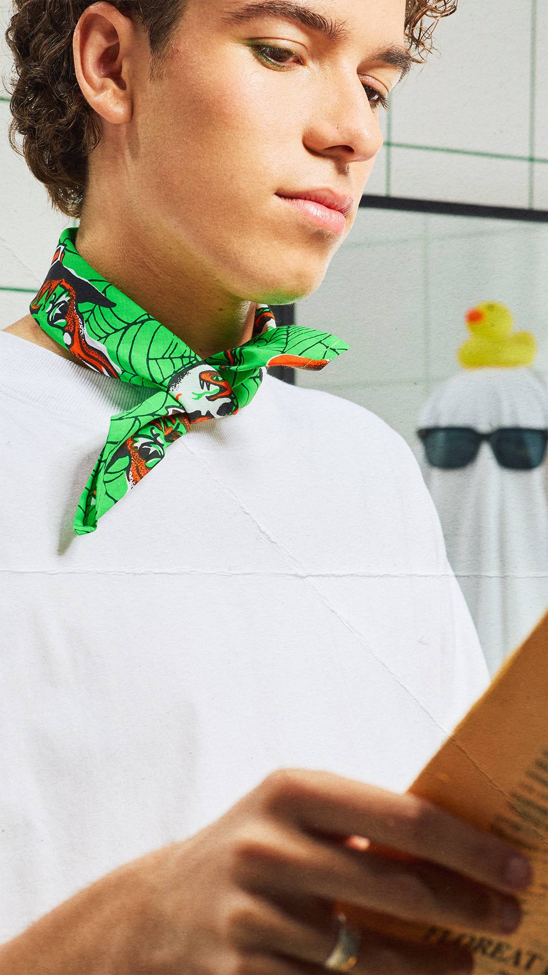 A close-up of a person wearing green eyeliner and a vibrant green Halloween knot-wrap tied around their neck like a scarf. In the background, a person disguised as a ghost with a white towel, sunglasses, and a rubber duck stands in the room.