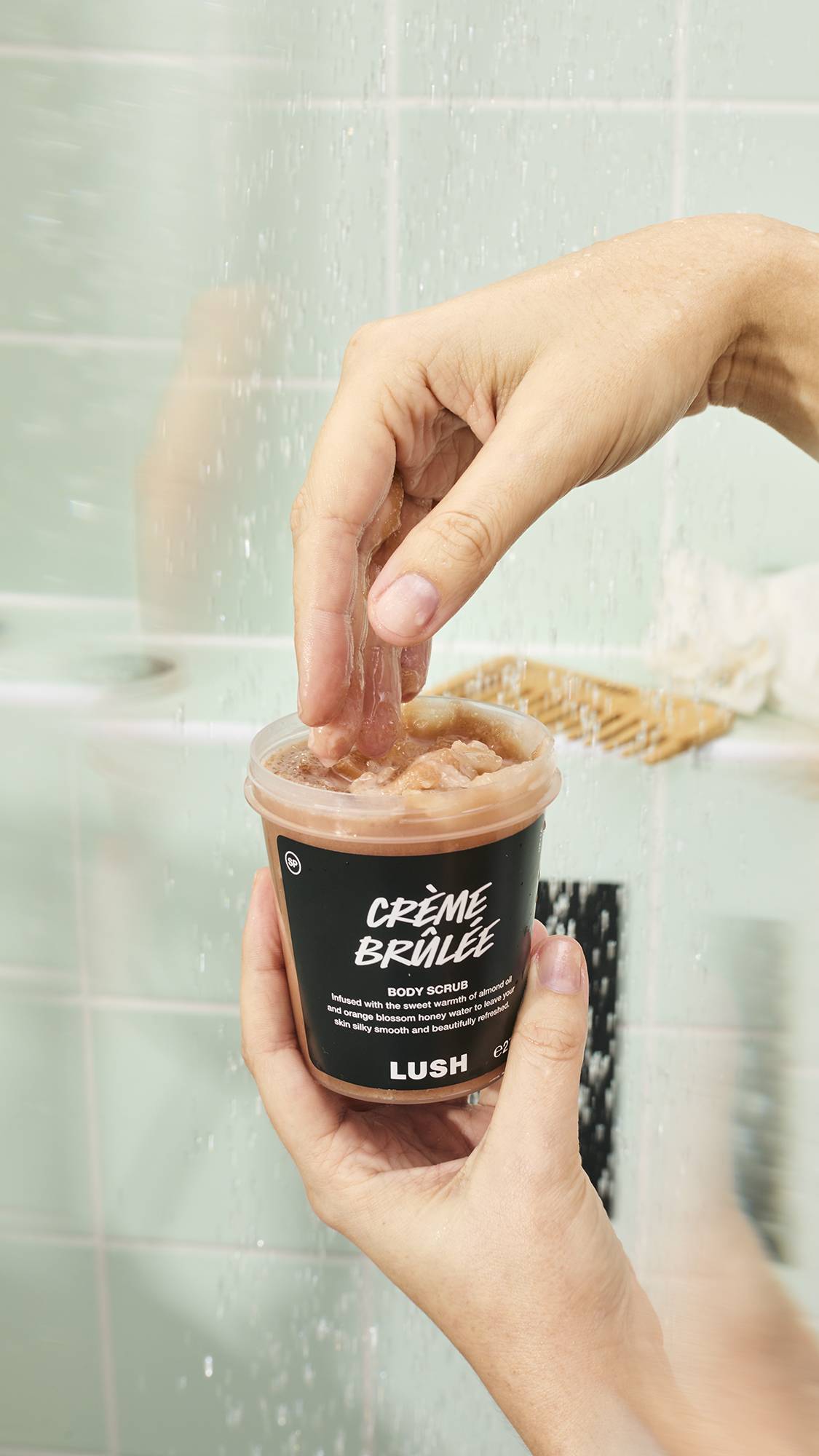 Wet hands hold an open pot of "Crème Brûlée" Body Scrub against a green tiled shower background. Fingers dip into the container to scoop out the product, which the label describes as being infused with "almond oil and orange blossom honey water".