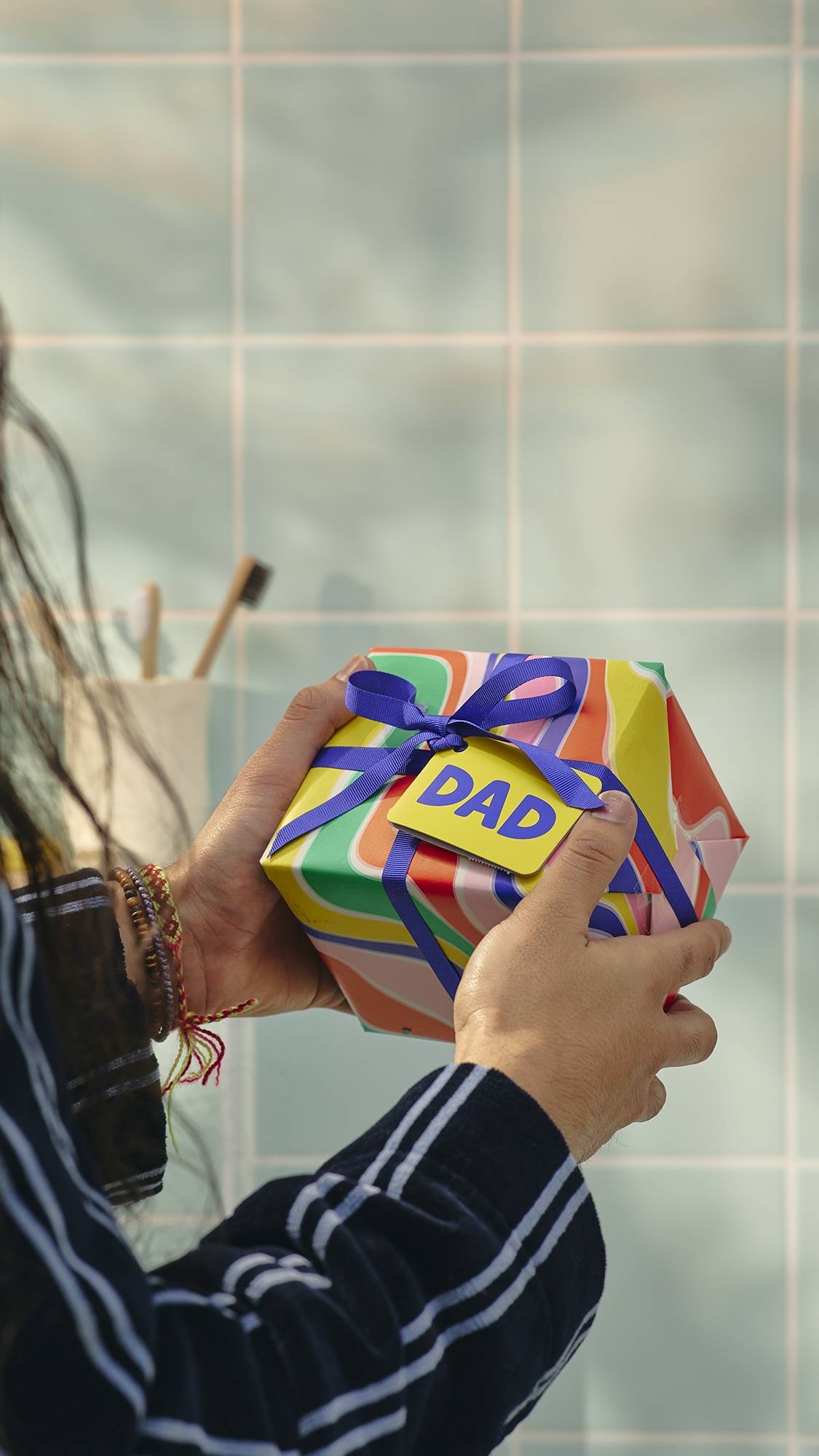 A close-up image of a model's hands holding a square gift wrapped in vibrant, swirling patterns of orange, yellow, and green. It is tied with a purple ribbon and a yellow tag that says 'DAD'. In the background are light blue bathroom tiles.