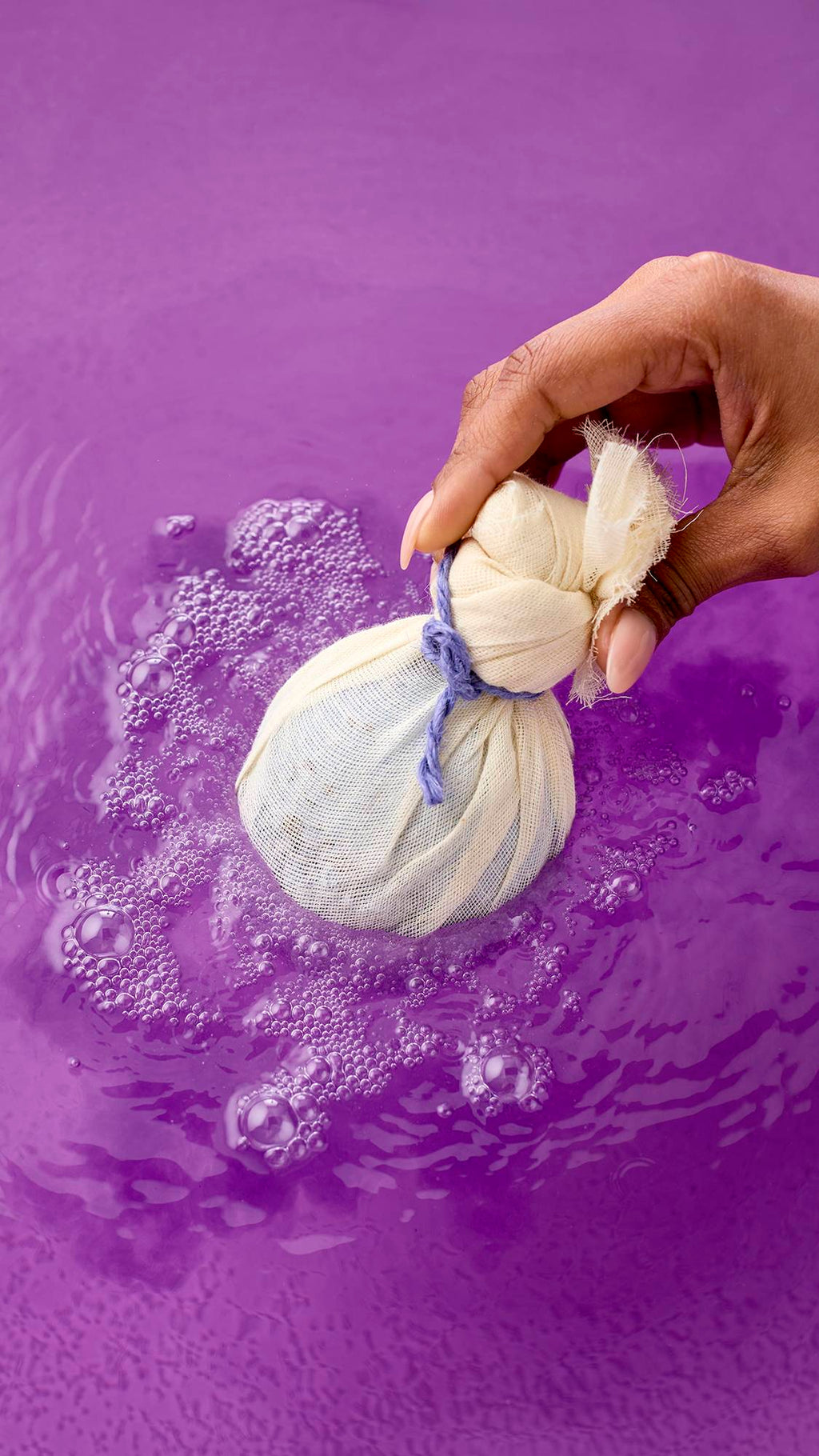 Deep Sleep bath bomb. The spherical bath bomb is wrapped in cream-colored muslin, tied with a purple cord, and half-submerged in water. The water reflects the vibrant purple, with ripples spreading across the surface from the product.