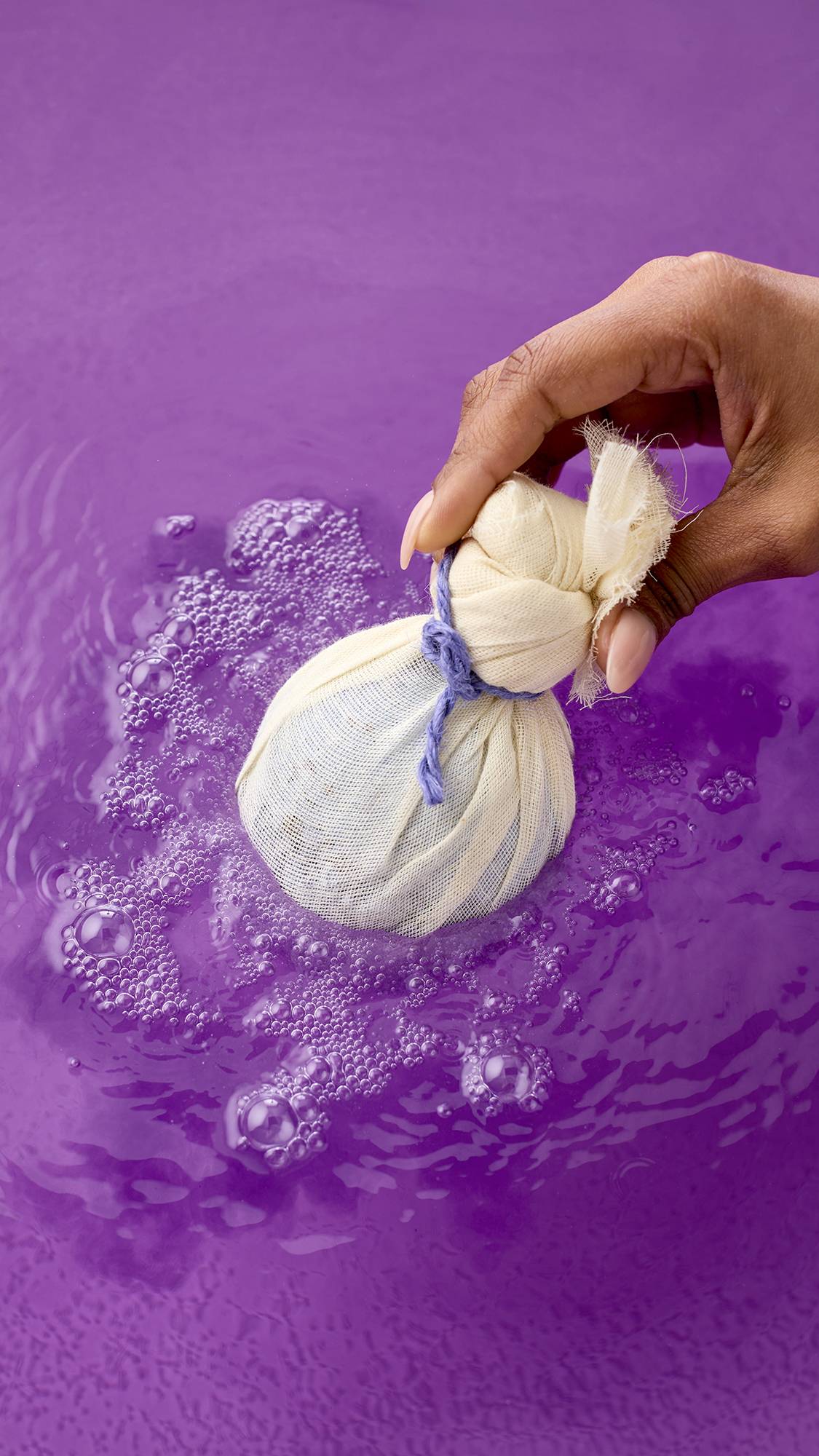 Deep Sleep bath bomb. The spherical bath bomb is wrapped in cream-colored muslin, tied with a purple cord, and half-submerged in water. The water reflects the vibrant purple, with ripples spreading across the surface from the product.