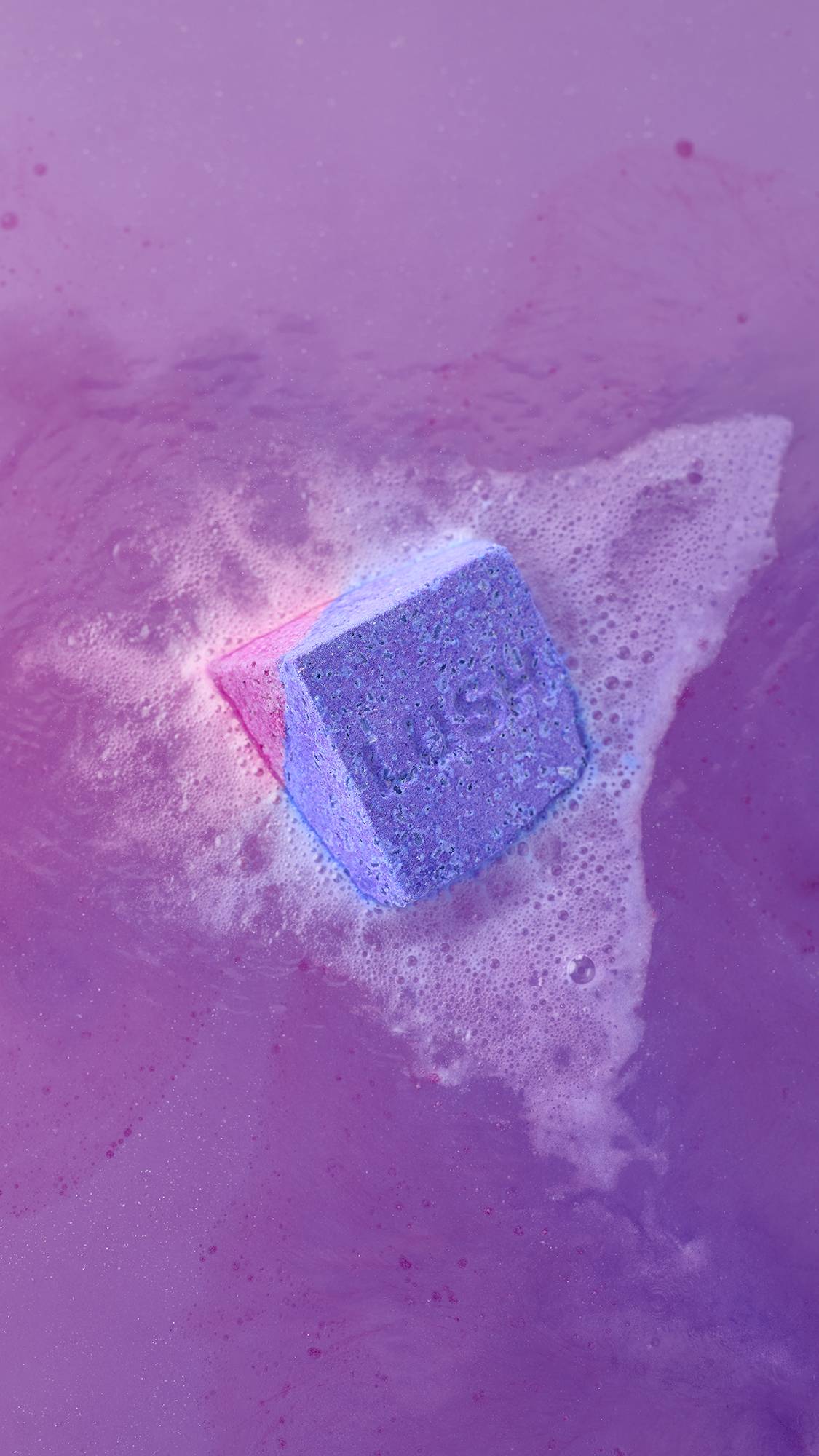 A top-down view of the Deep sleep Salt Cube bath bomb fizzing in vibrant purple water. The pink and purple cube is surrounded by a thick, frothy cloud of white bubbles and foam.