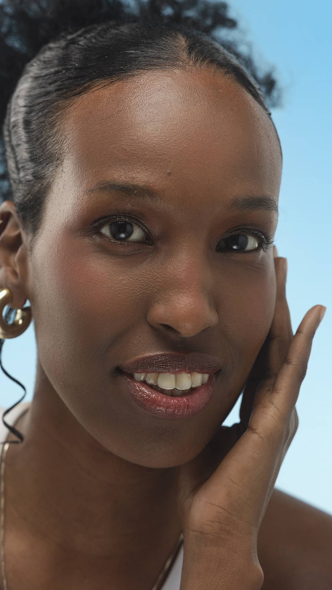 A person with dark skin smiles, touching their face. They wear a subtle mauve-brown lip color and gold earrings, against a blue background, likely finished with Emotional Brilliance ~ Finishing Powder.