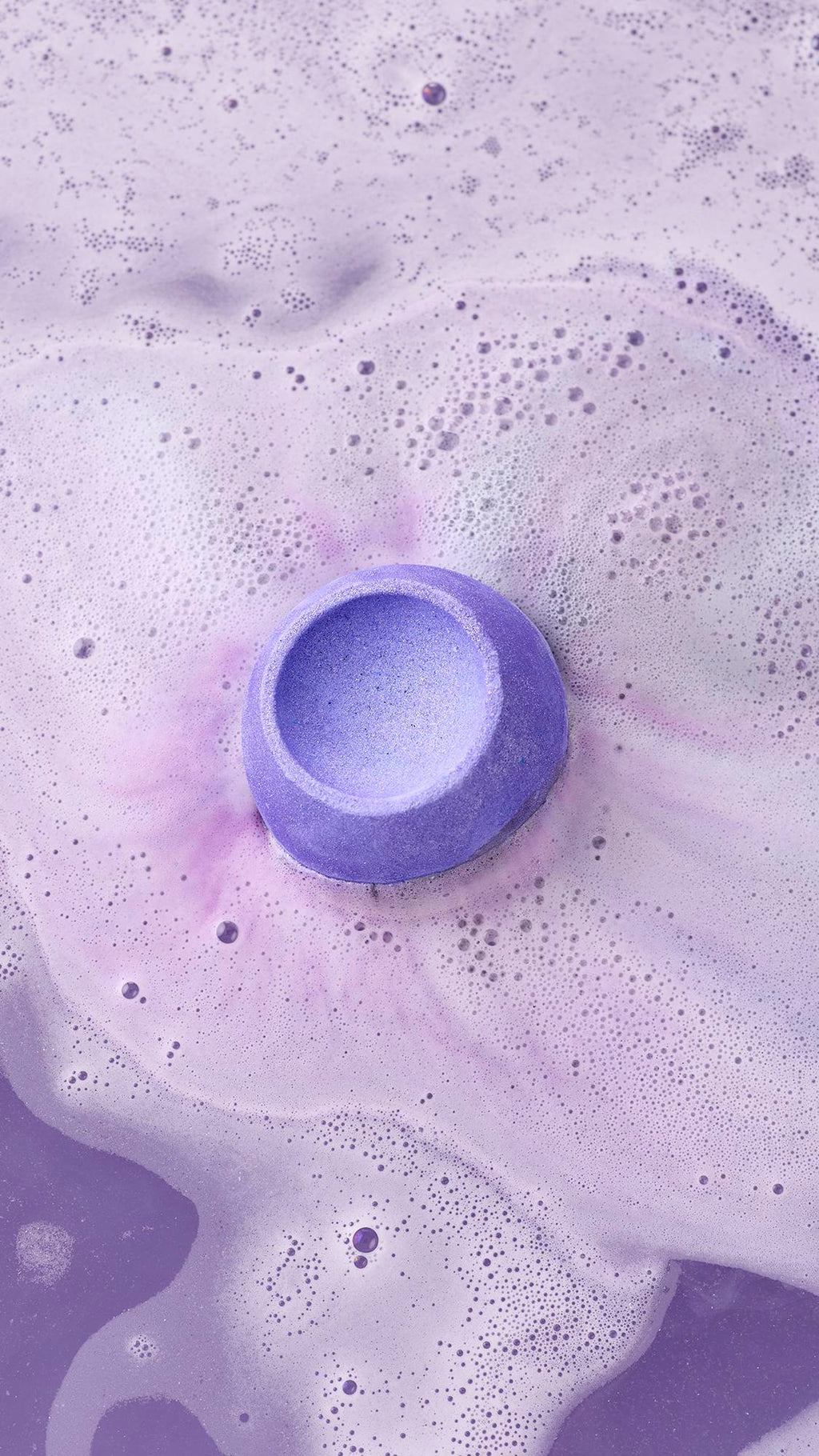A vibrant purple Lush "Goddess" bath bomb fizzes in lilac-coloured water, creating a thick layer of white and pale purple foam. The bath bomb has a bowl-shaped indentation on top and is surrounded by delicate bubbles on the water's surface.