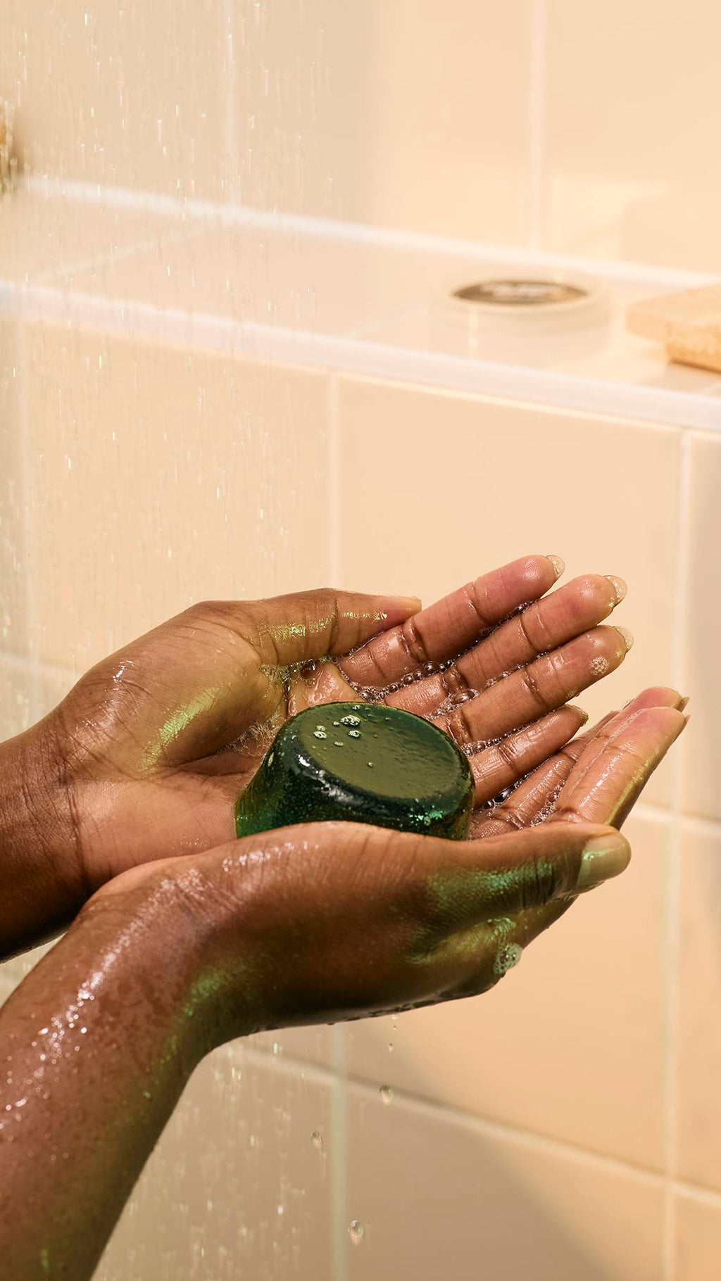 A model's hands hold a dark green, translucent "The Good Hour" shower jelly in their cupped hands under shower water. The jelly has a smooth, dome shape, and small, shimmering bubbles form on its surface as the water hits.