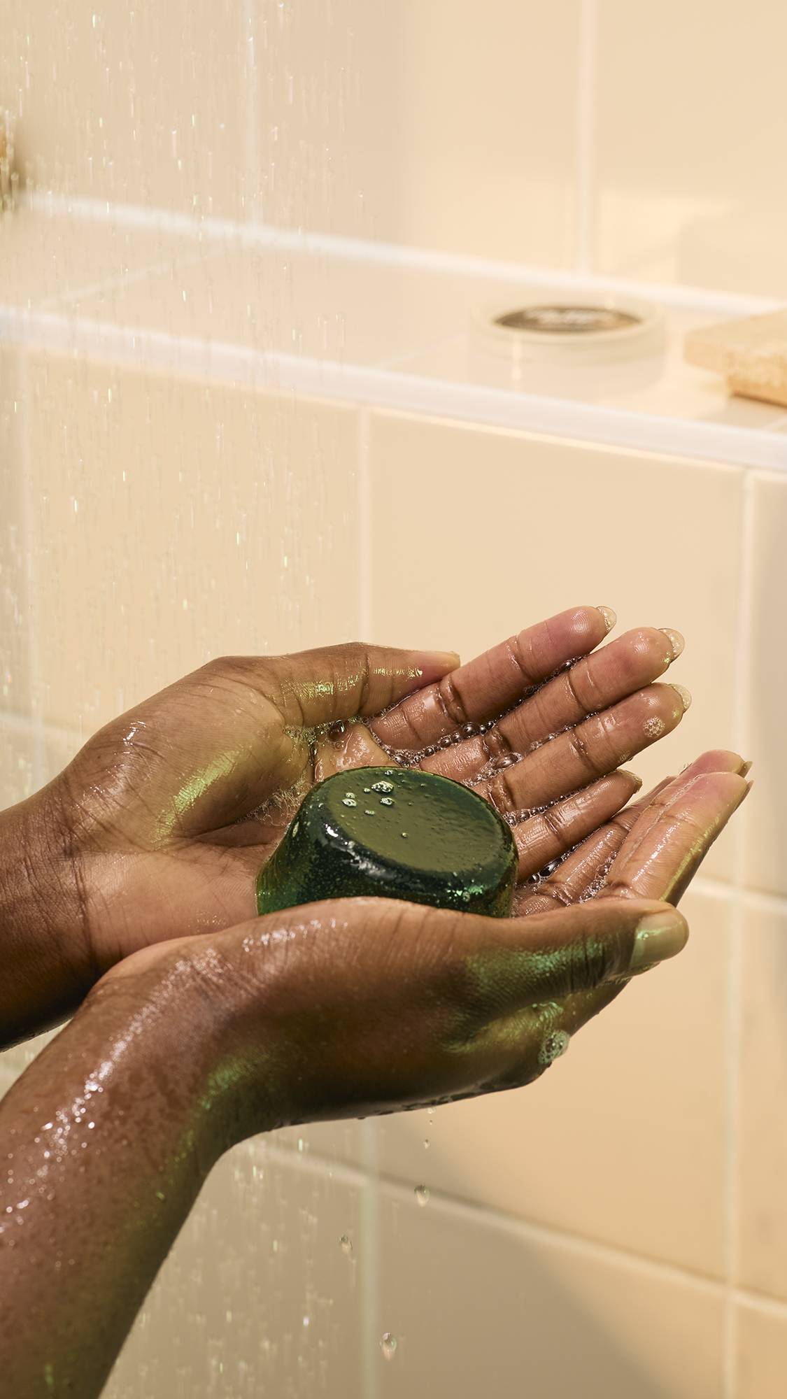 A model's hands hold a dark green, translucent "The Good Hour" shower jelly in their cupped hands under shower water. The jelly has a smooth, dome shape, and small, shimmering bubbles form on its surface as the water hits.