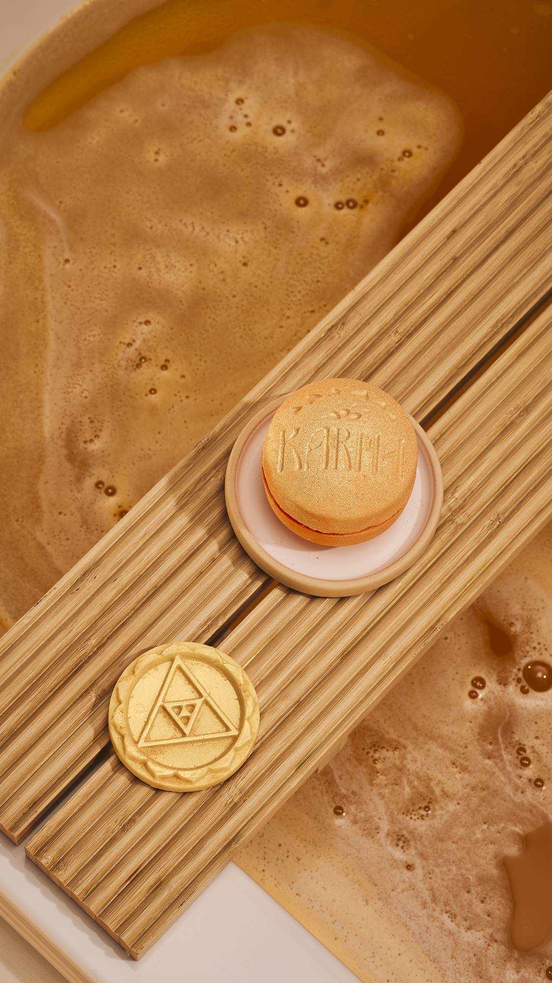 An orange, macaron-shaped 'Karma' bath bomb rests on a ceramic dish. Beside it on a wooden bath tray is the pale-yellow, circular middle layer embossed with a triangle design. Below, the bathwater is a deep amber colour with a thick layer of foam.