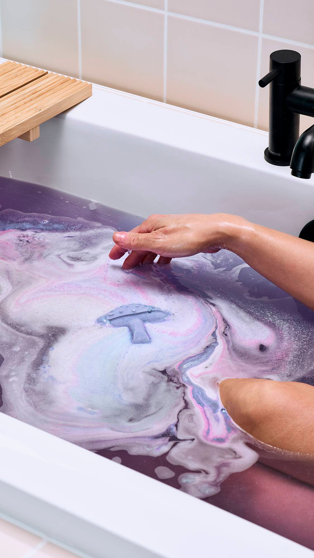 The image shows the purple "Magik Mushroom" floating in a bath of deep lilac water. It is surrounded by swirls of white, pink, and pale blue foam as it dissolves. A model's arm reaches towards the floating mushroom from the side.